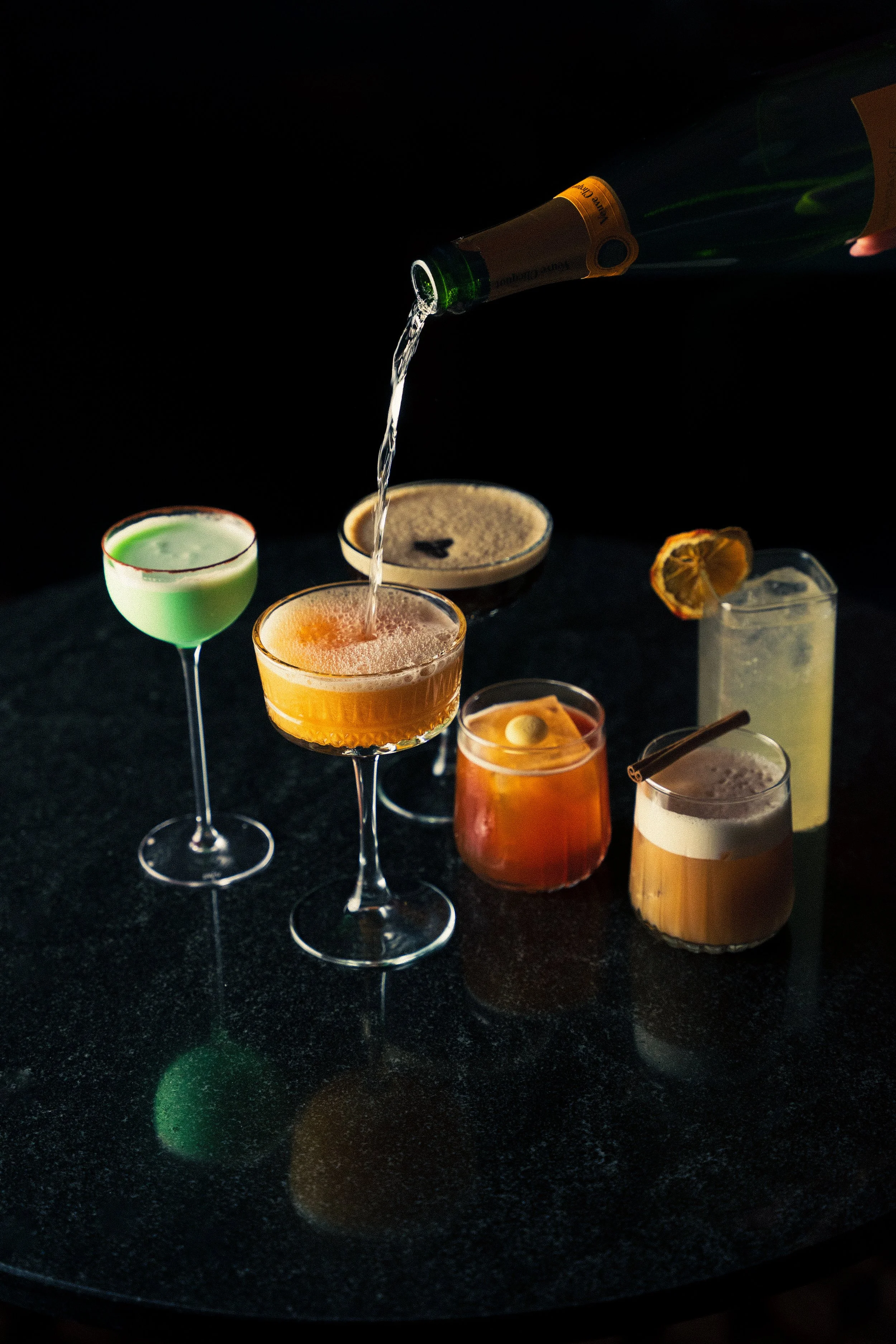 A row of colorful cocktails on a black surface, with someone pouring a drink into the middle glass.
