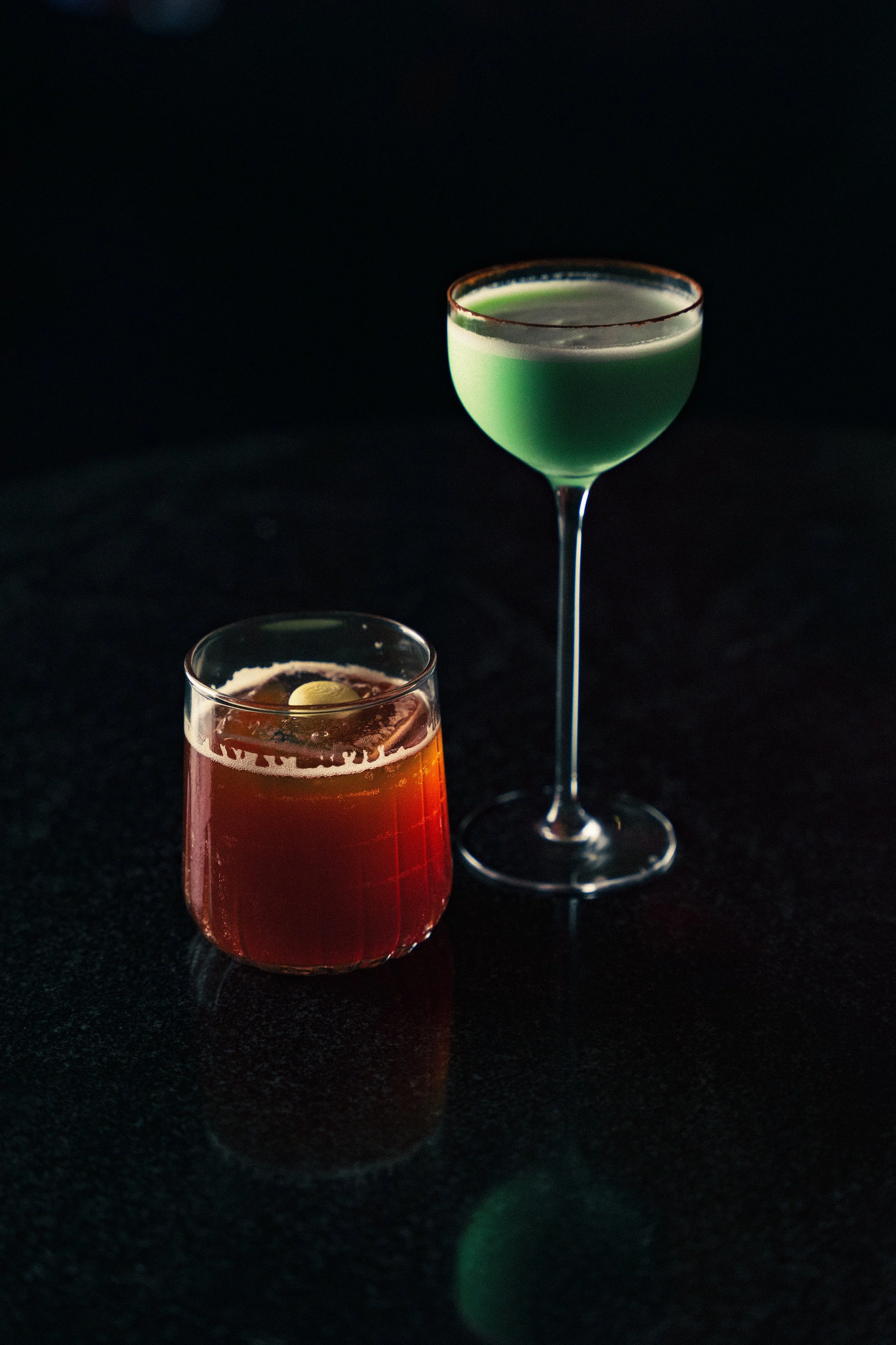 Two cocktails on a dark surface, one in a short glass with an olive garnish and one in a tall martini glass with a green liquid.