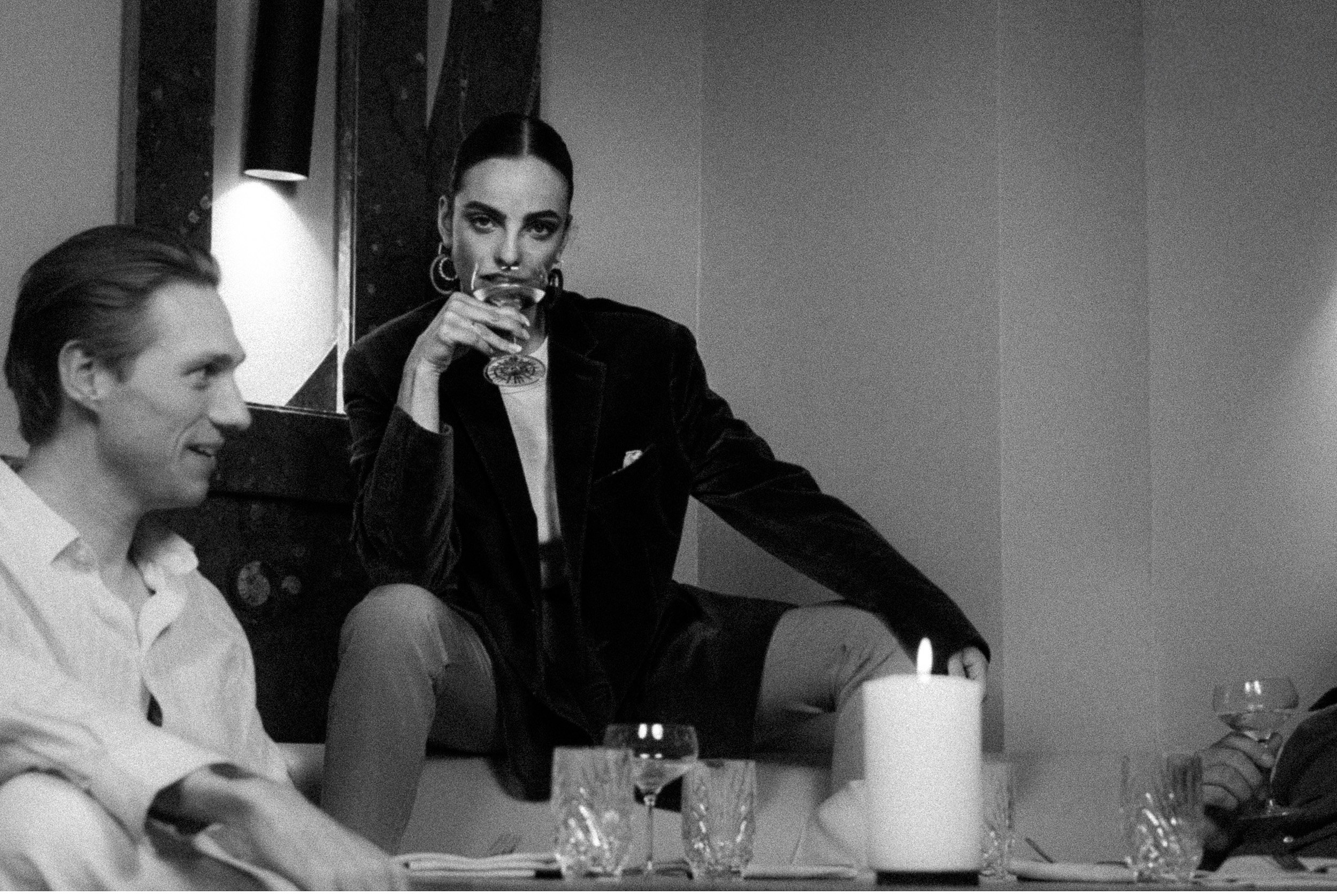 Black and white photo of three people at a dining table, with a woman holding a glass and sitting on a ledge, and two men seated, one smiling and the other holding a glass.