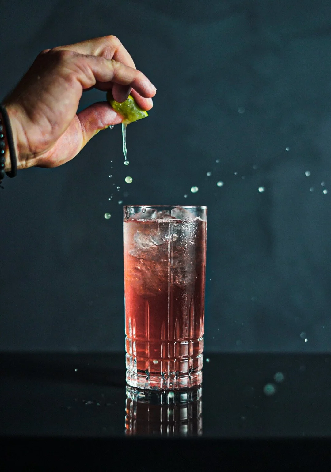 A hand squeezing a lime into a tall glass of red-colored drink with ice, against a dark background.