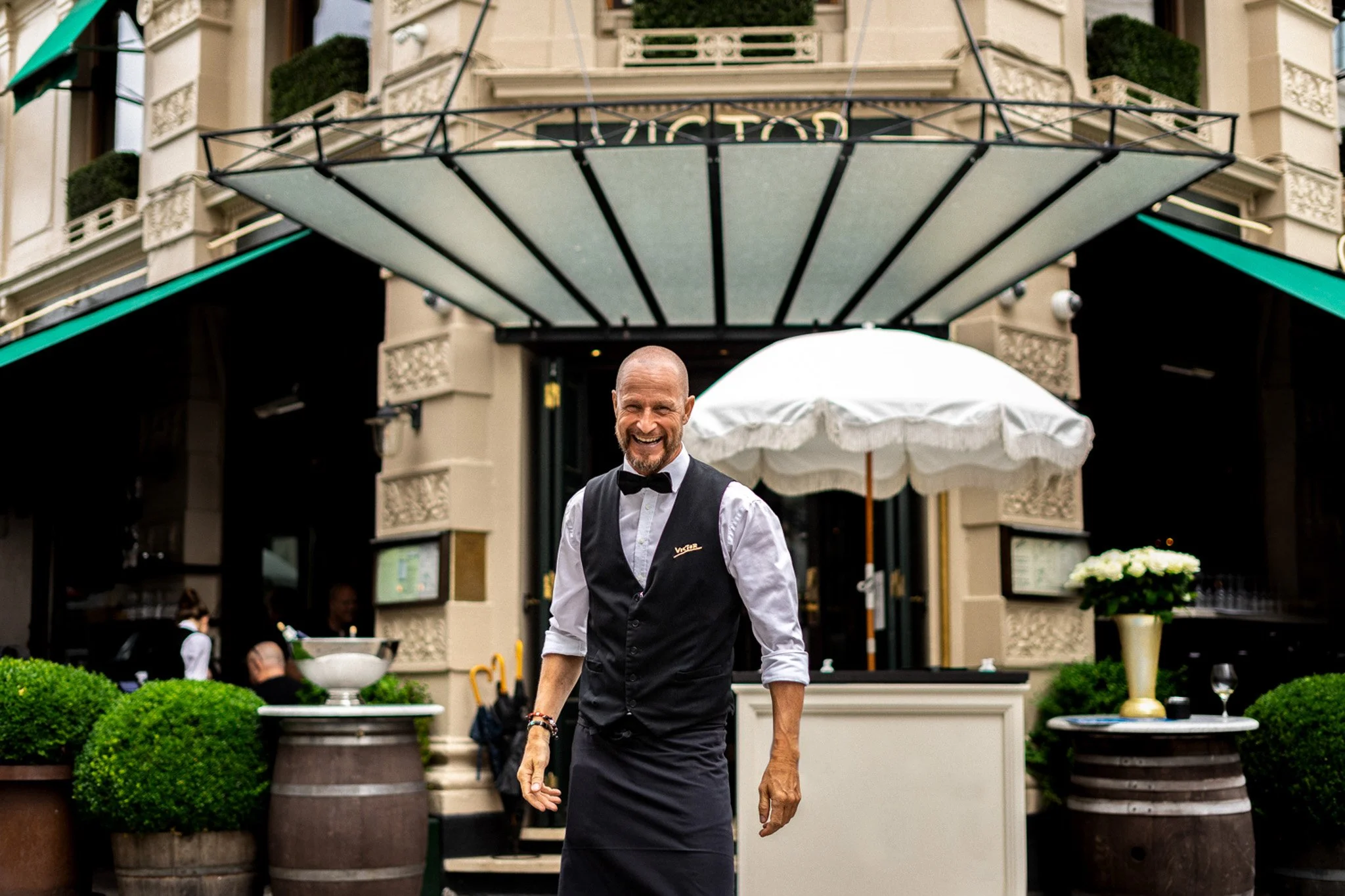 A smiling man dressed as a waiter in a black apron and bow tie stands outside a building entrance, with a white umbrella and decorative plants in pots around him.