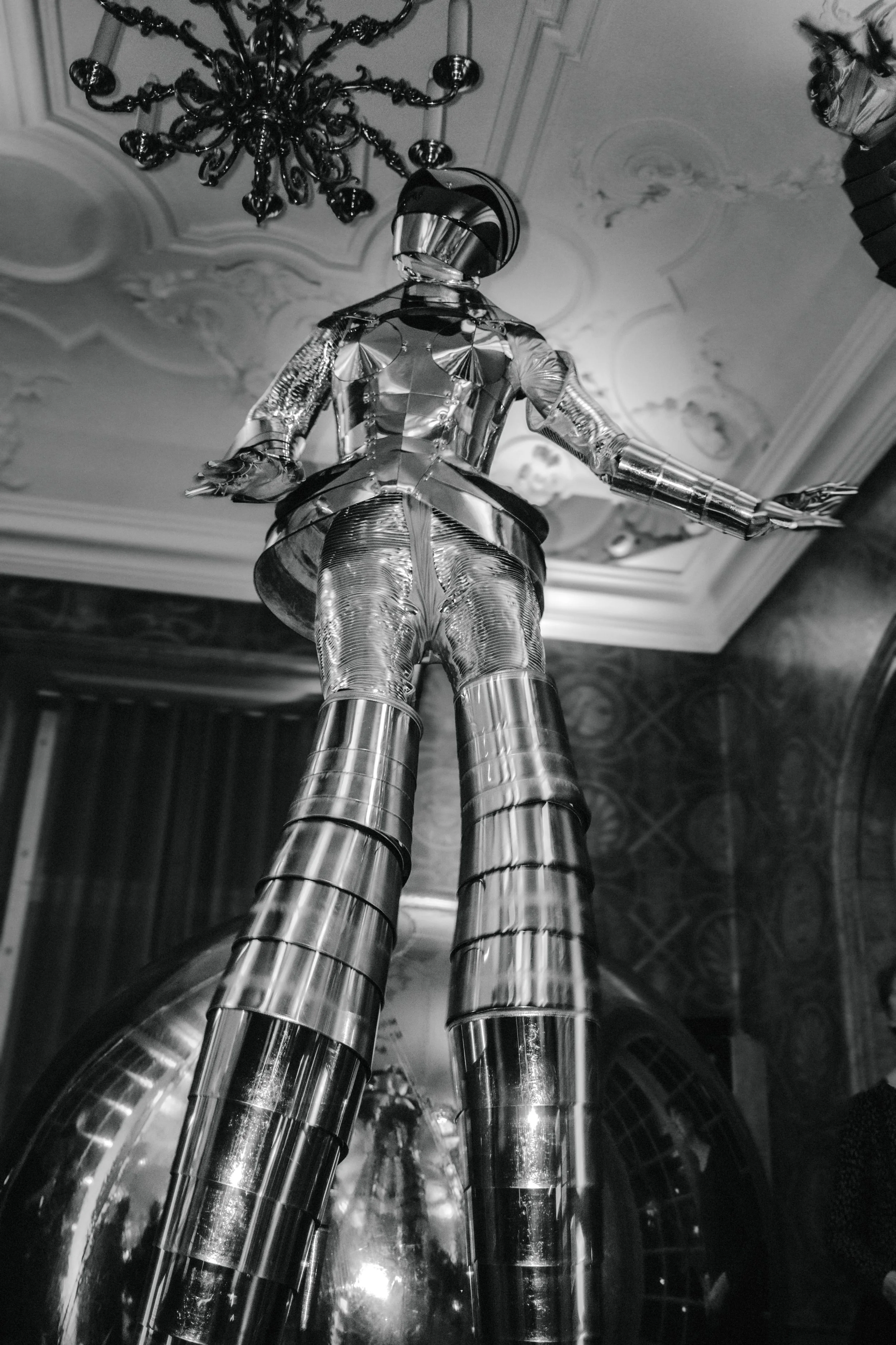 A tall, reflective, metallic sculpture of a woman in a form-fitting outfit stands in an elegant room with ornate ceiling details and a chandelier, viewed from a low angle.