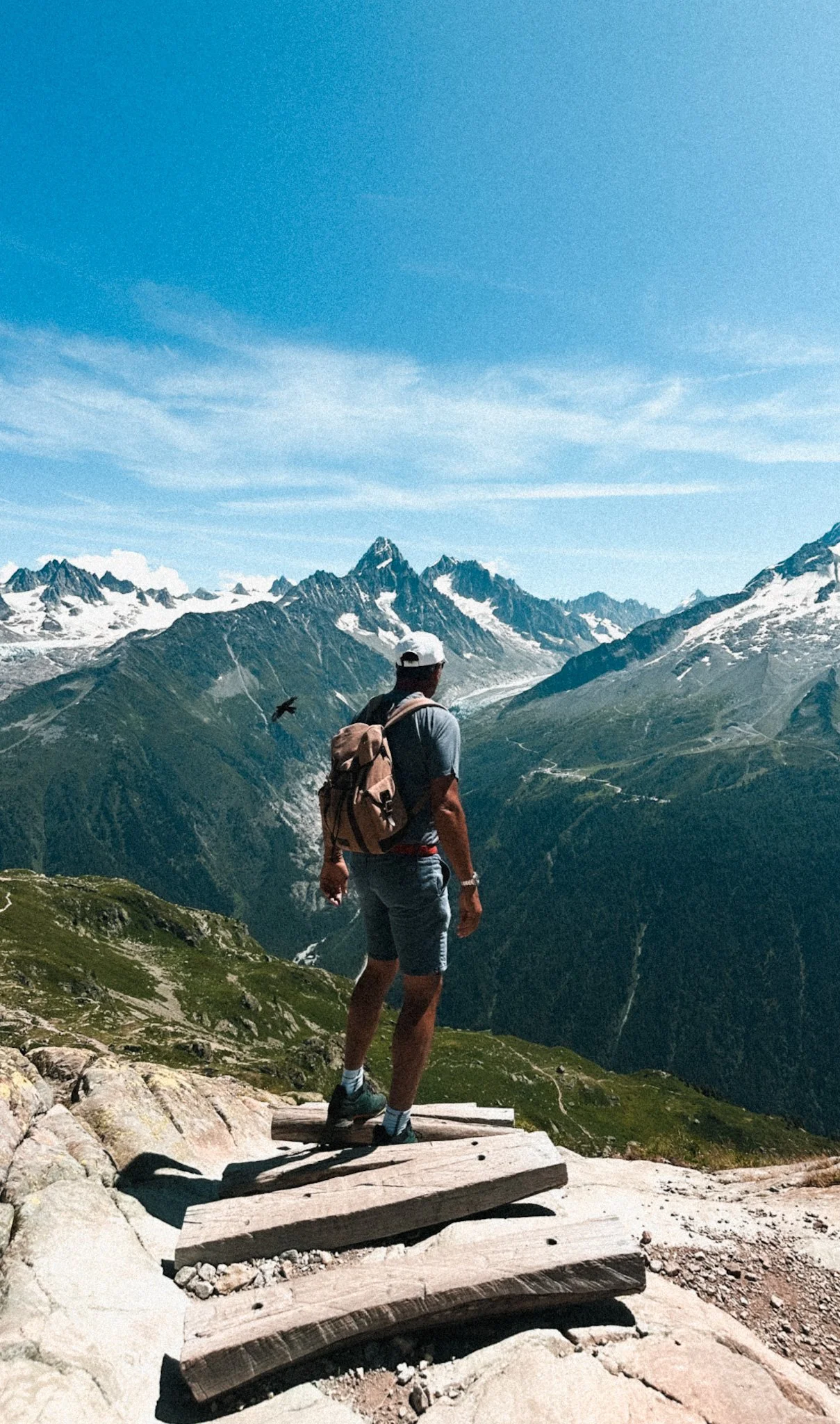 Man with a backpack standing on a wooden platform overlooking green mountains and snow-capped peaks under a blue sky with clouds.