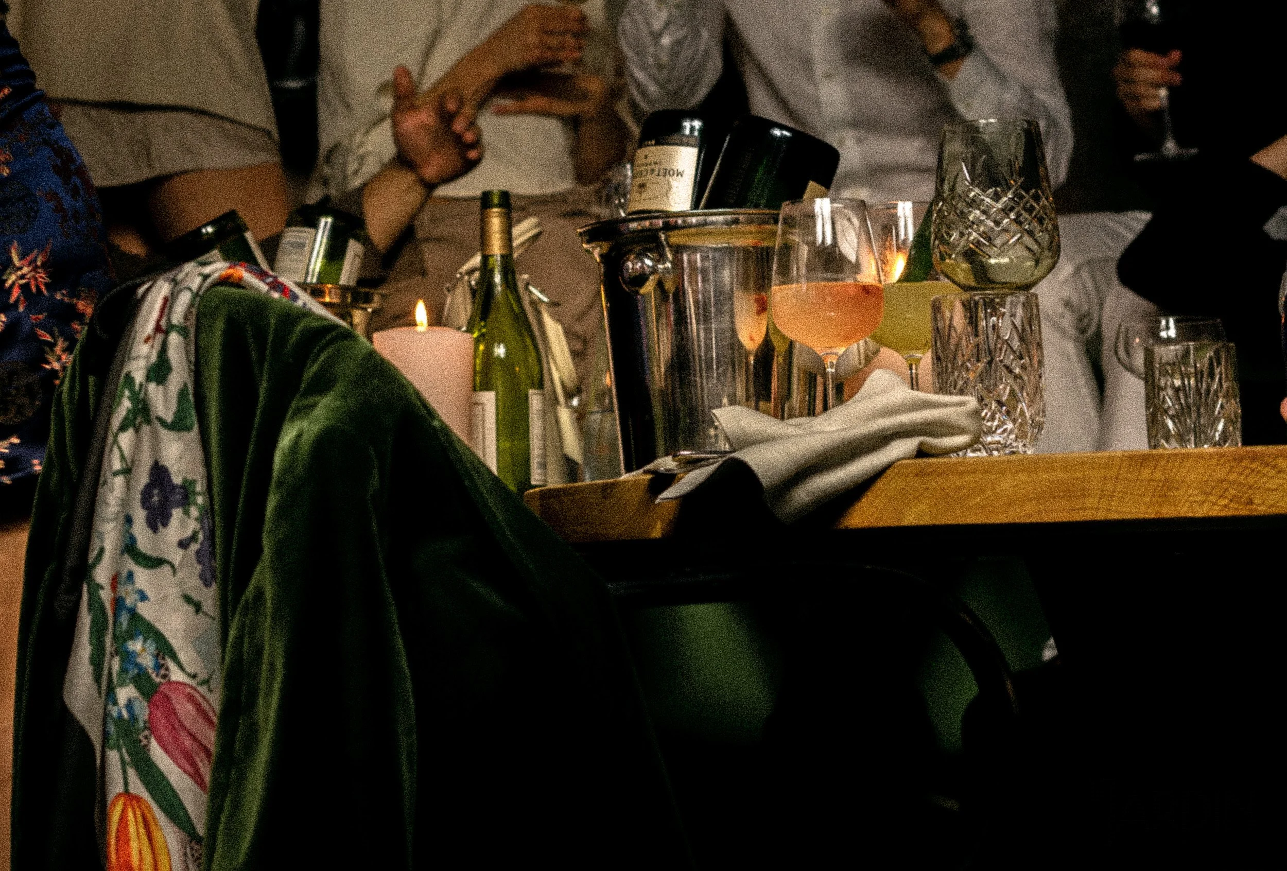 A table with candles, wine bottles, glasses of wine, and various drinking glasses at a social gathering, with people in the background.