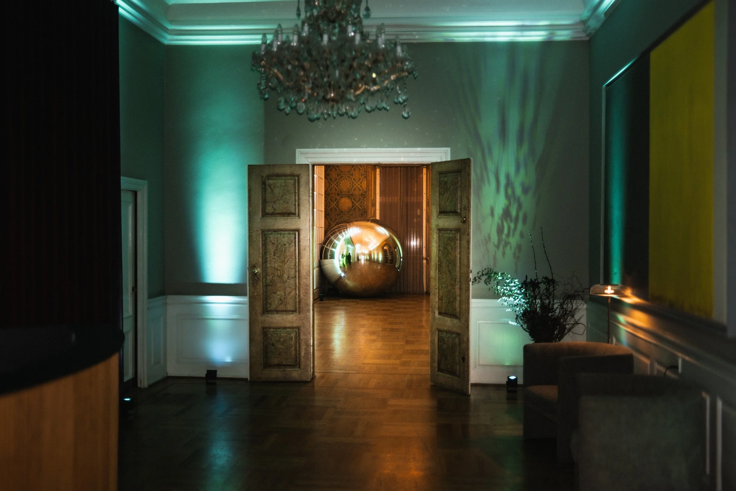 An elegant interior room with a chandelier, painted walls with lighting, and double wooden doors opening into a room with a reflective metallic sphere on the floor.
