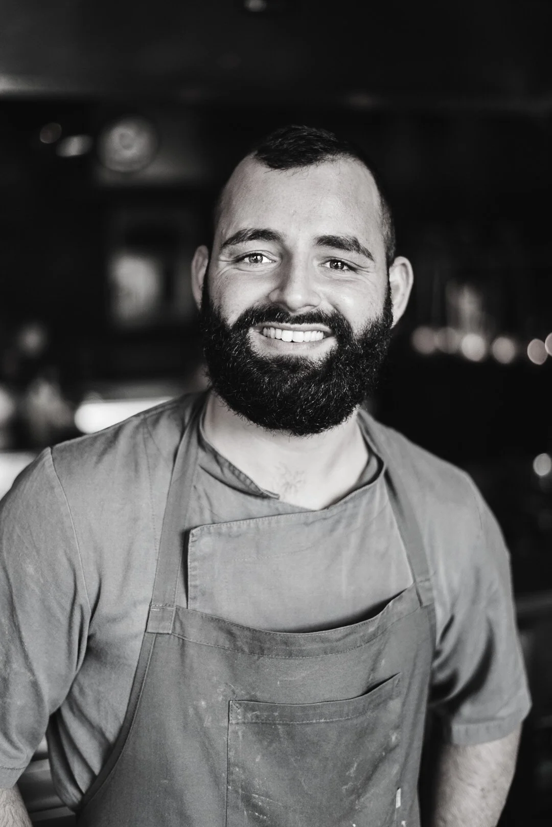 A smiling man with a beard wearing a dark apron in a professional kitchen.