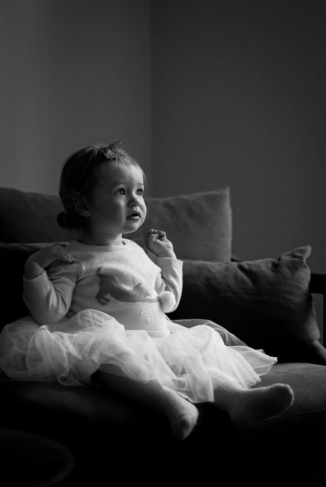 A young girl with a sweater featuring a horse and a tutu-like skirt, sitting on a couch and looking at something with a surprised or curious expression.