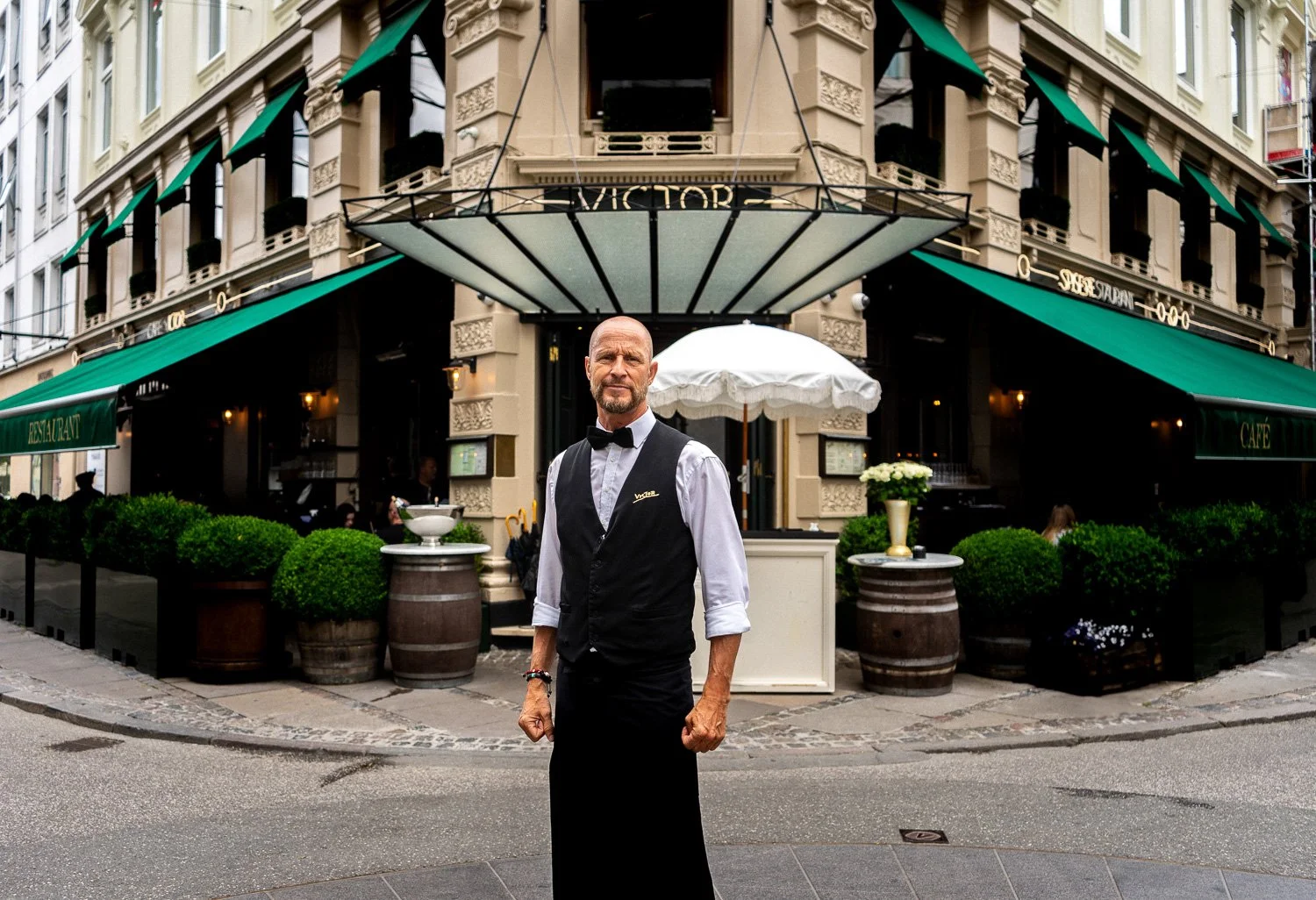 A waiter in a black vest and white shirt with rolled-up sleeves and a bowtie stands outside a restaurant with green awnings. The restaurant has classic architecture with ornate details, and the street is paved with cobblestones.