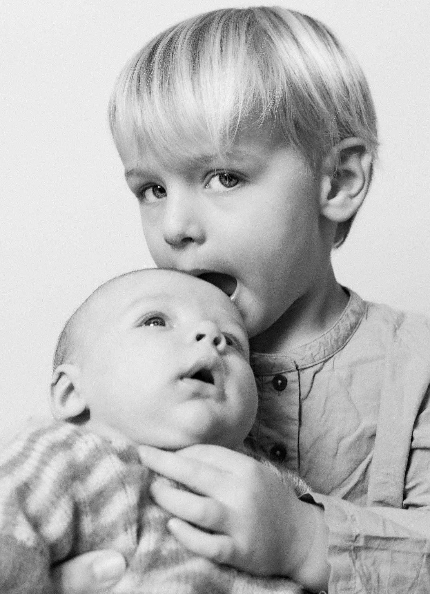 A young boy with light hair holds a baby close, near his face, as the baby looks upward. The photo is in black and white.