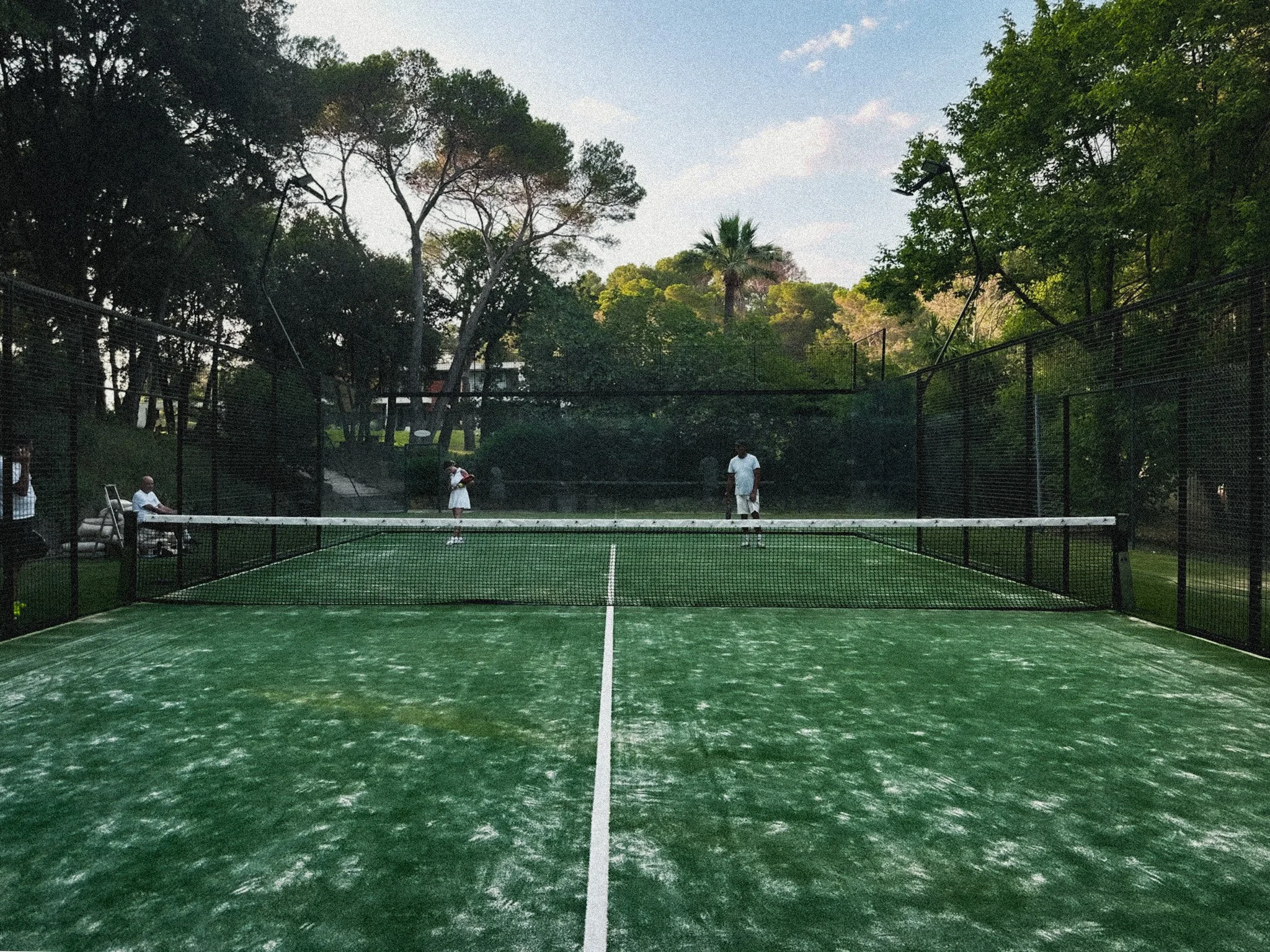 A fenced outdoor pickleball court with four players and a person sitting on a bench, surrounded by trees under a partly cloudy sky.