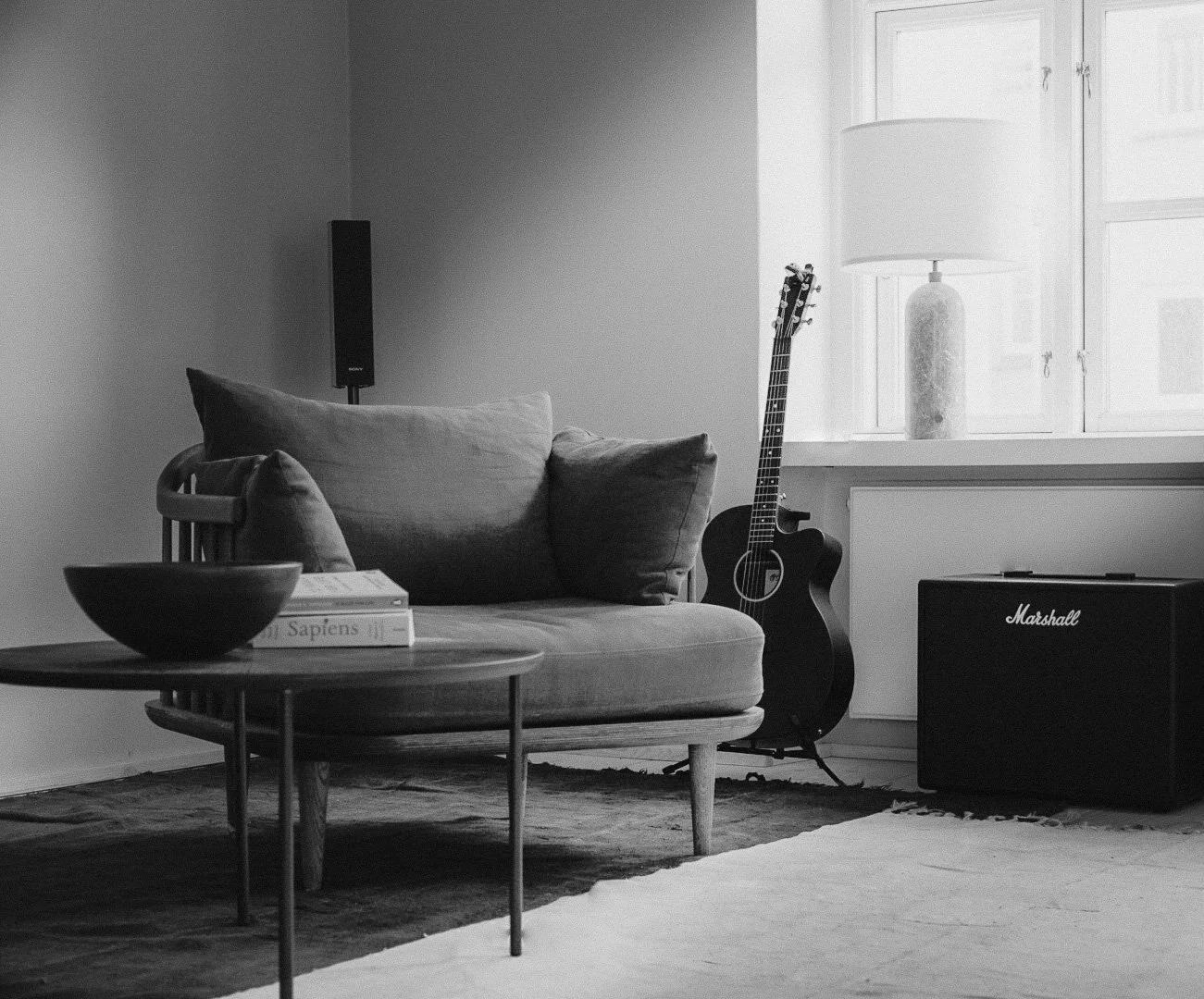 A living room corner with a sofa, a round wooden coffee table, books, a ceramic bowl, a guitar, a Marshall amplifier, a window with a large lampshade, and a speaker, all in black and white.
