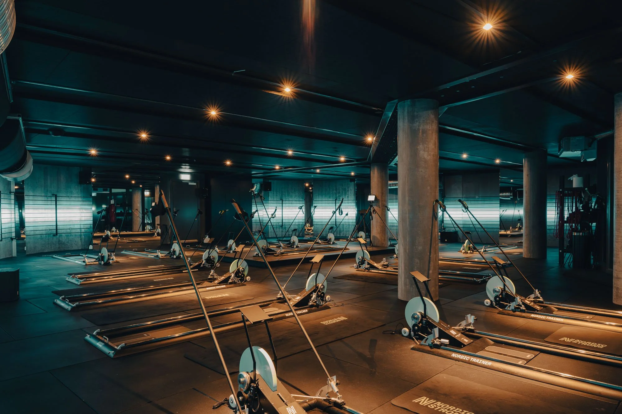 Empty indoor gym with multiple rowing machines arranged in rows, dim lighting, and some illuminated horizontal light strips on the walls.