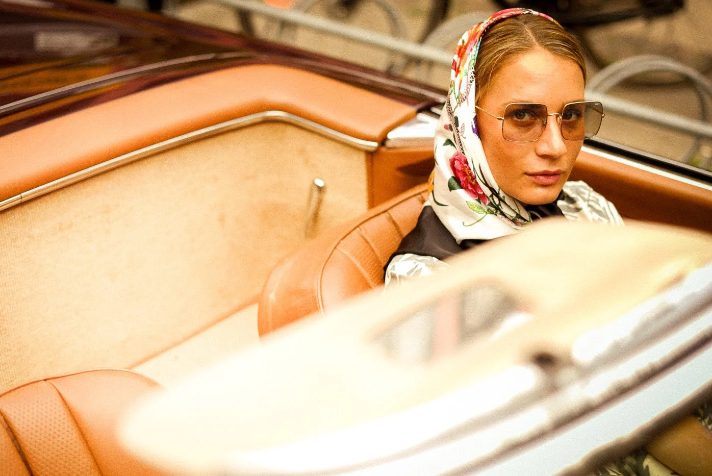 A woman sitting in a vintage car, wearing sunglasses, a floral headscarf, and stylish clothing, with a neutral expression.