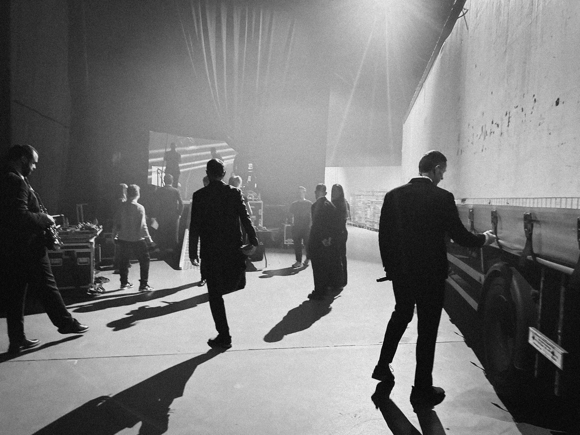 Black and white photo of people walking backstage at an event, with stage lights and equipment in the background.
