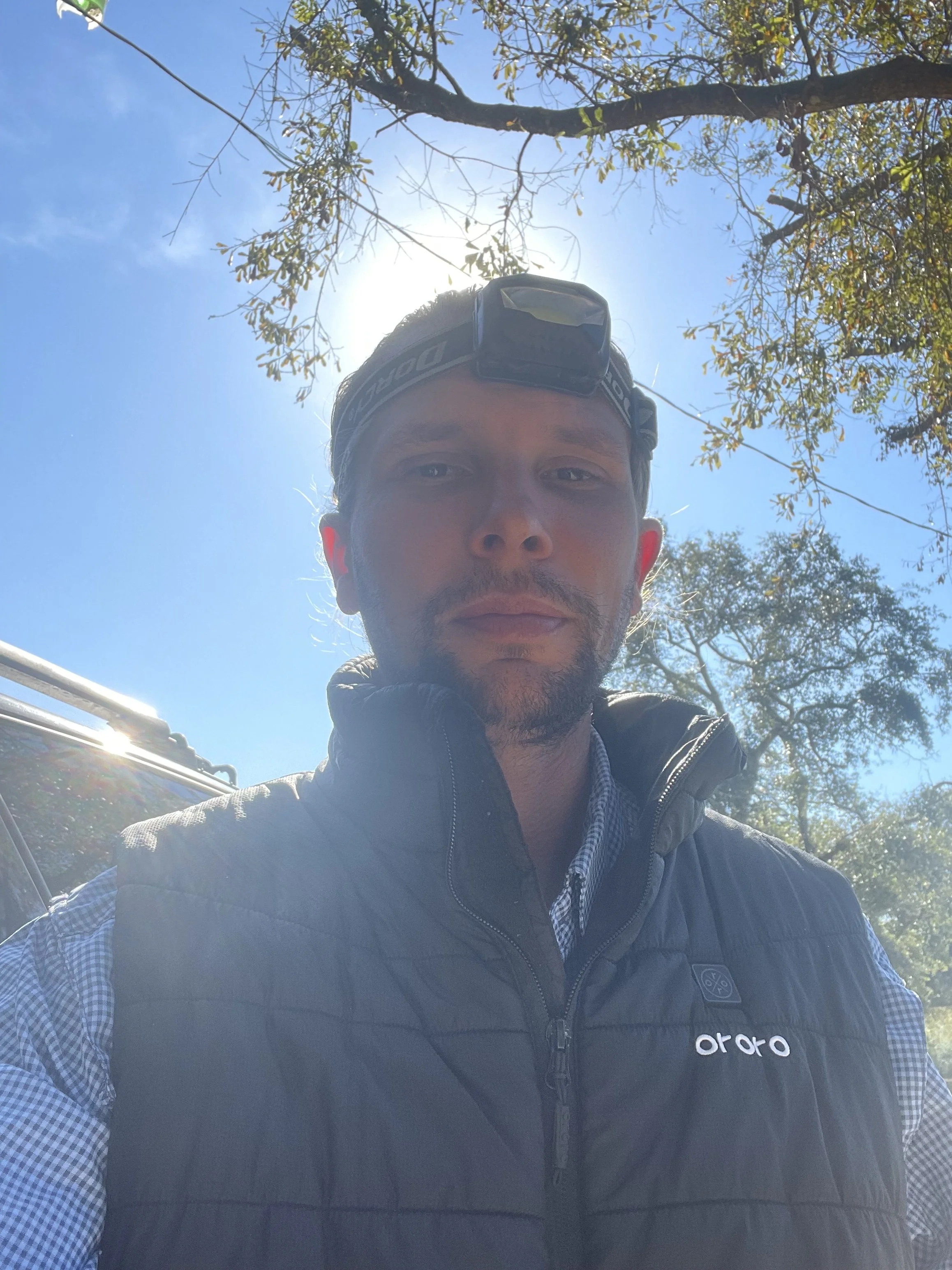 A man wearing a black vest, checkered shirt, and headlamp takes a selfie outdoors on a sunny day with trees and a clear blue sky in the background.