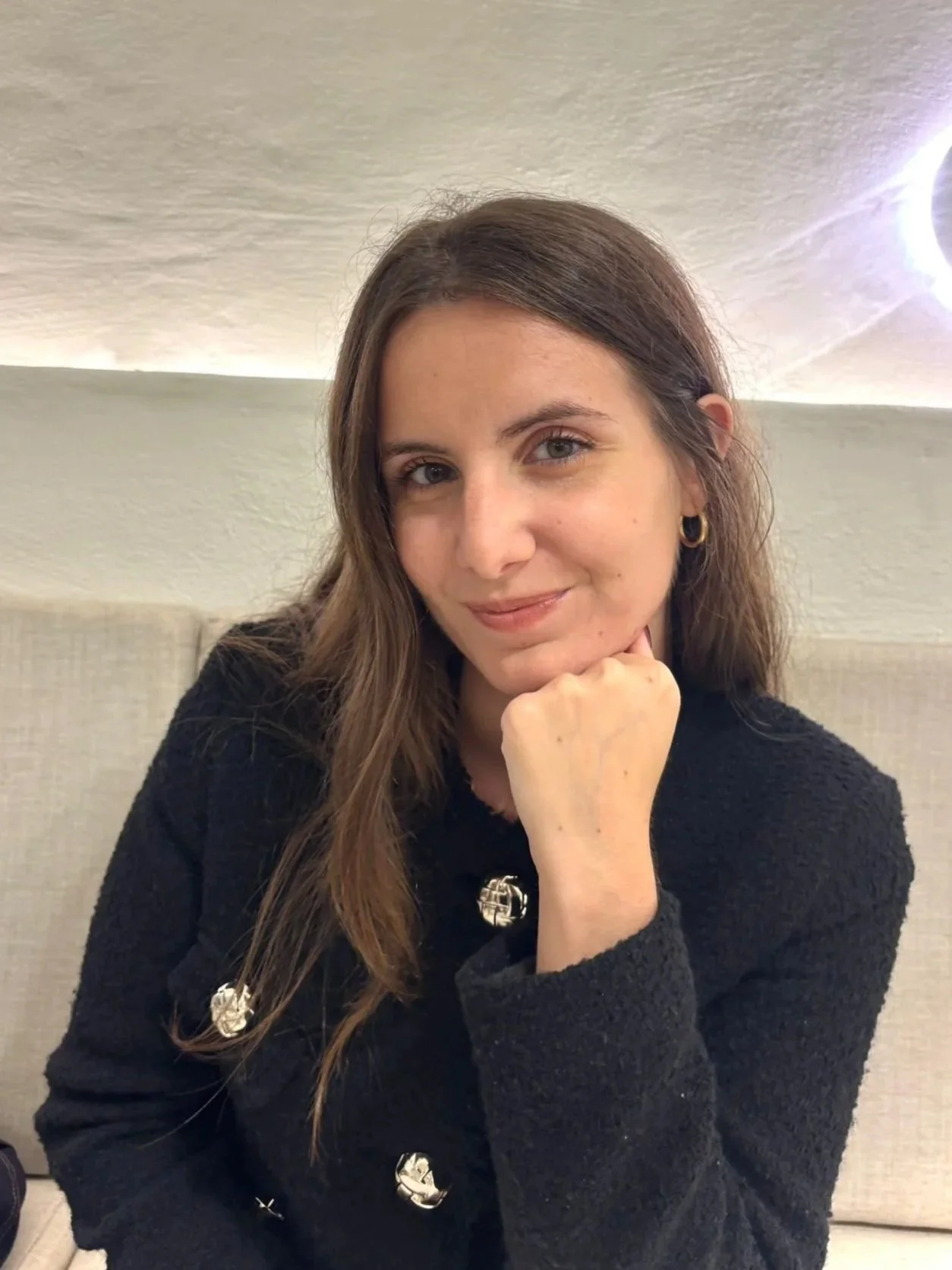 Una mujer con cabello largo y castaño, usando un suéter negro con botones dorados, está sonriendo y apoyando la mandíbula con su mano, en un ambiente iluminado.