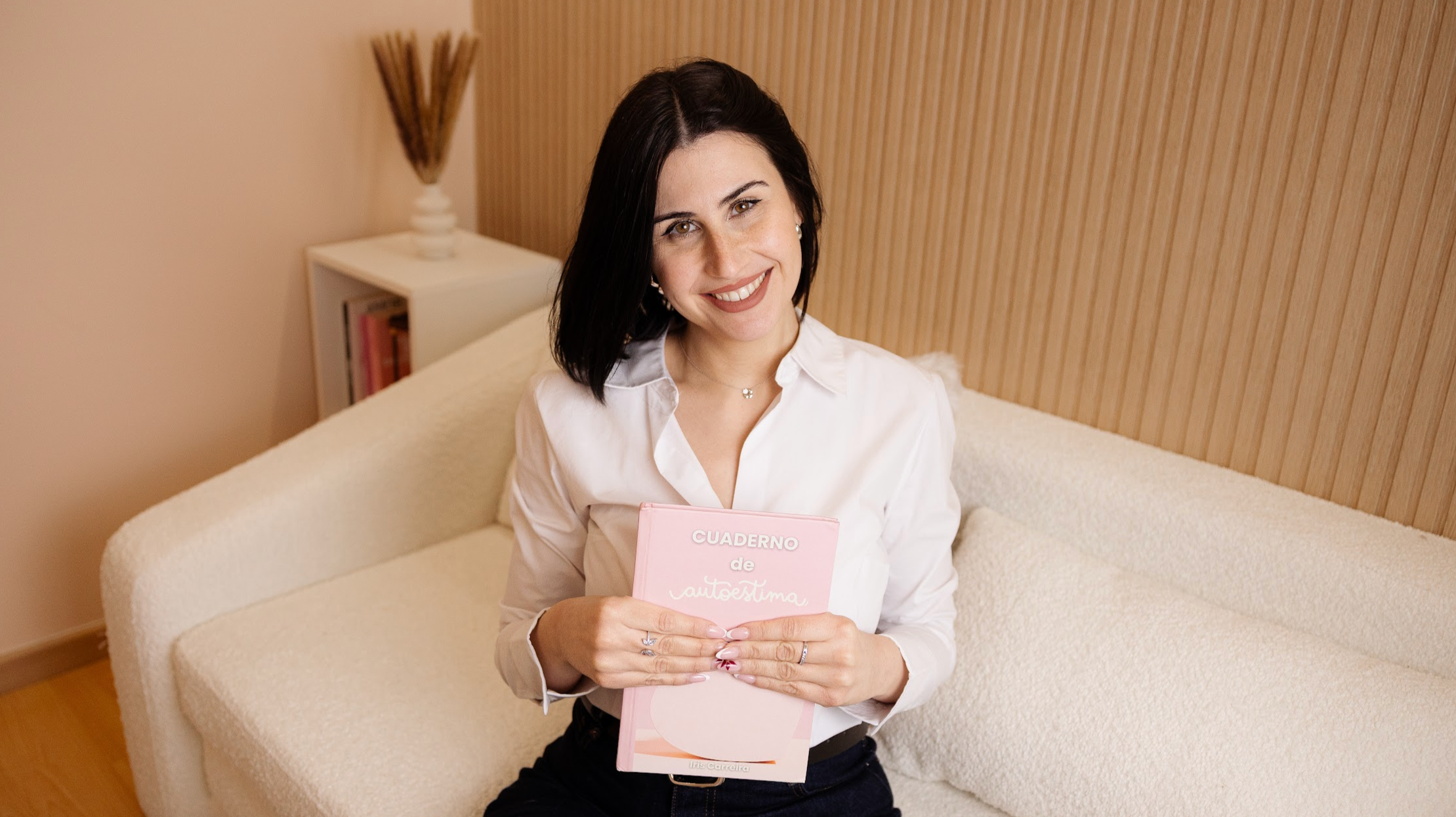 Mujer sonriendo sosteniendo un cuaderno rosa en un ambiente hogareño.