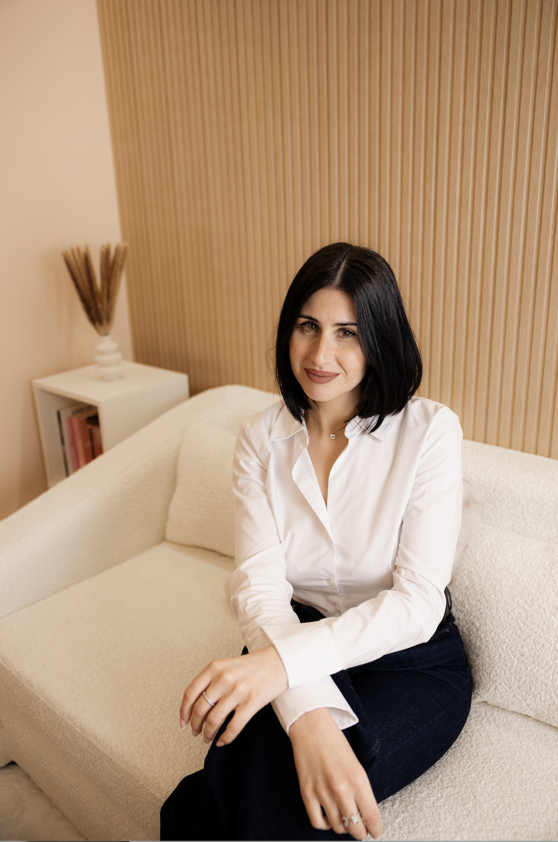 Mujer sentada en un sofá en una sala con pared de madera, vestida con blusa blanca y pantalones oscuros.