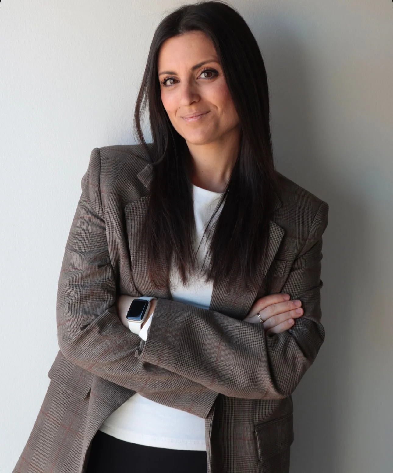 Retrato de una mujer con cabello oscuro, vistiendo un blazer a cuadros y camiseta blanca, sonriendo, con brazos cruzados y usando un reloj inteligente en la muñeca izquierda.