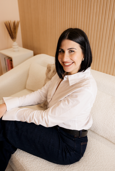 Mujer sonriendo, sentada en un sofá en un ambiente acogedor con pared de madera y una mesa con libro y decoraciones en segundo plano.