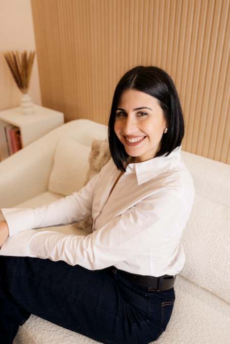 Mujer sonriendo sentada en un sofá con fondo de pared de madera y una mesa con libro y decoraciones.