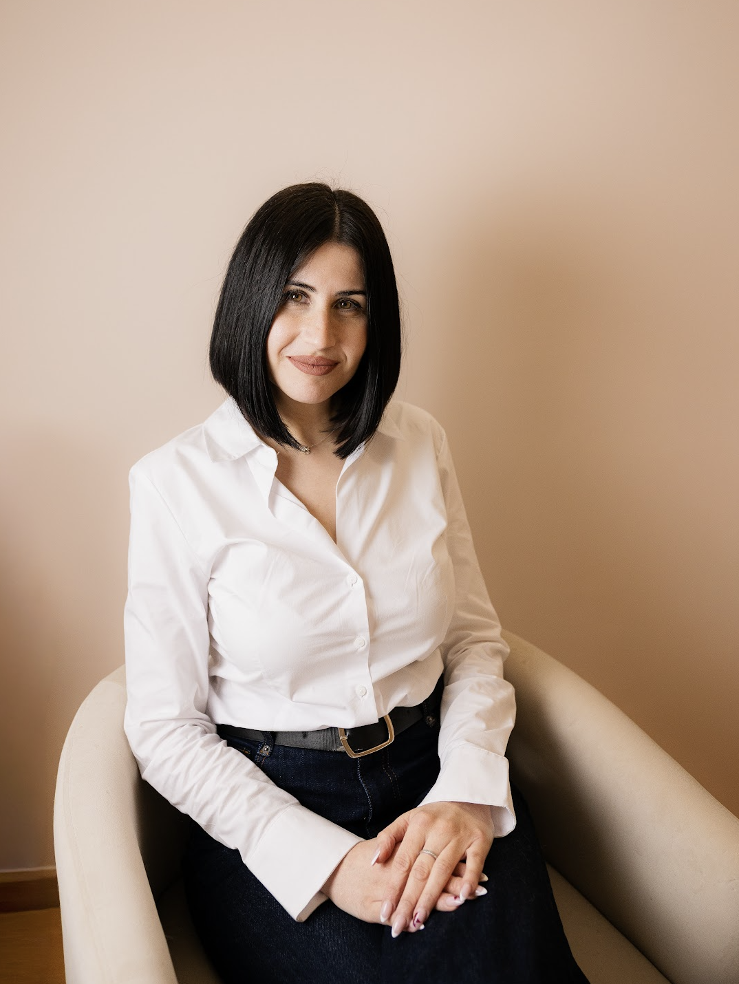 Mujer con cabello negro y corto, con blusa blanca y falda negra, sentada en un sillón beige, sonriendo.