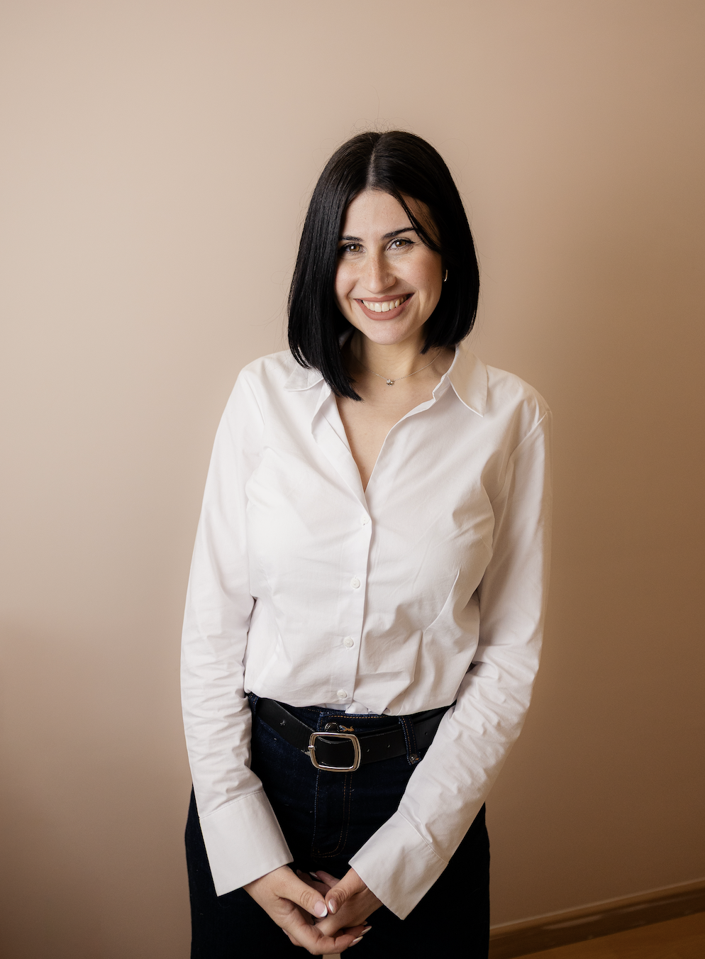 Mujer joven con cabello negro, usando blusa blanca y pantalón oscuro, sonriendo en un fondo beige.