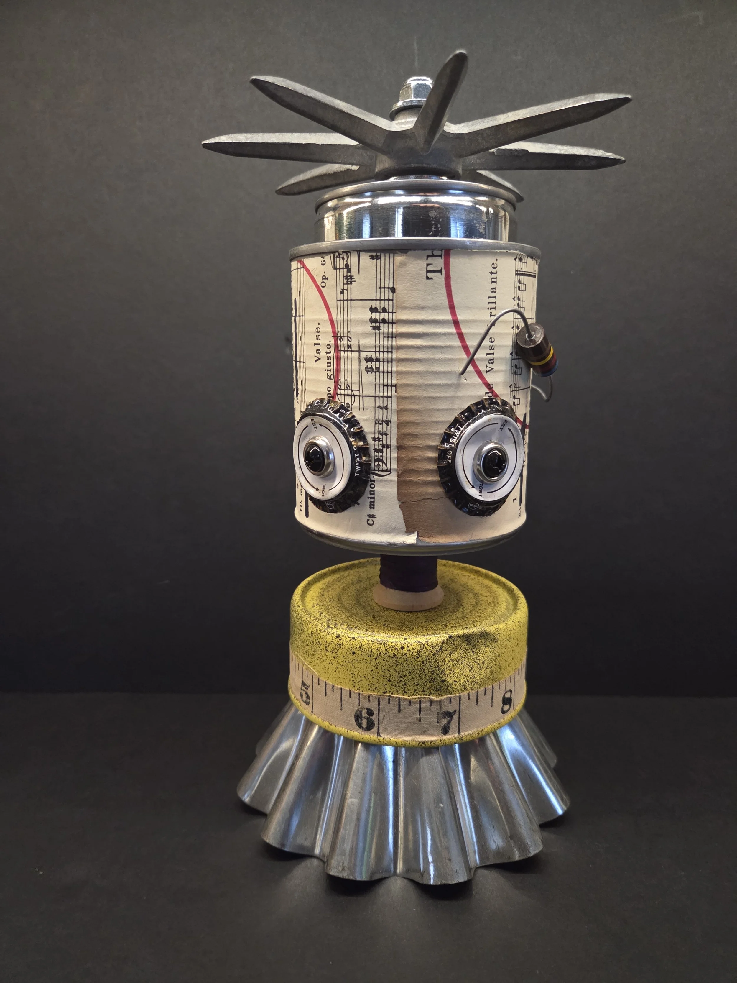 A robot sculpture made from recycled metal parts, with a can for a body, metal washers for eyes, a yellow measuring tape for a neck, a fluted metal base, and a propeller on top.