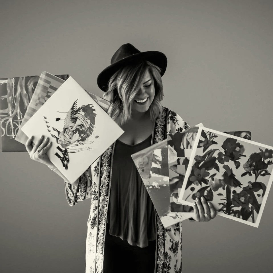 Woman with a hat smiling and holding several art prints with floral and animal designs.