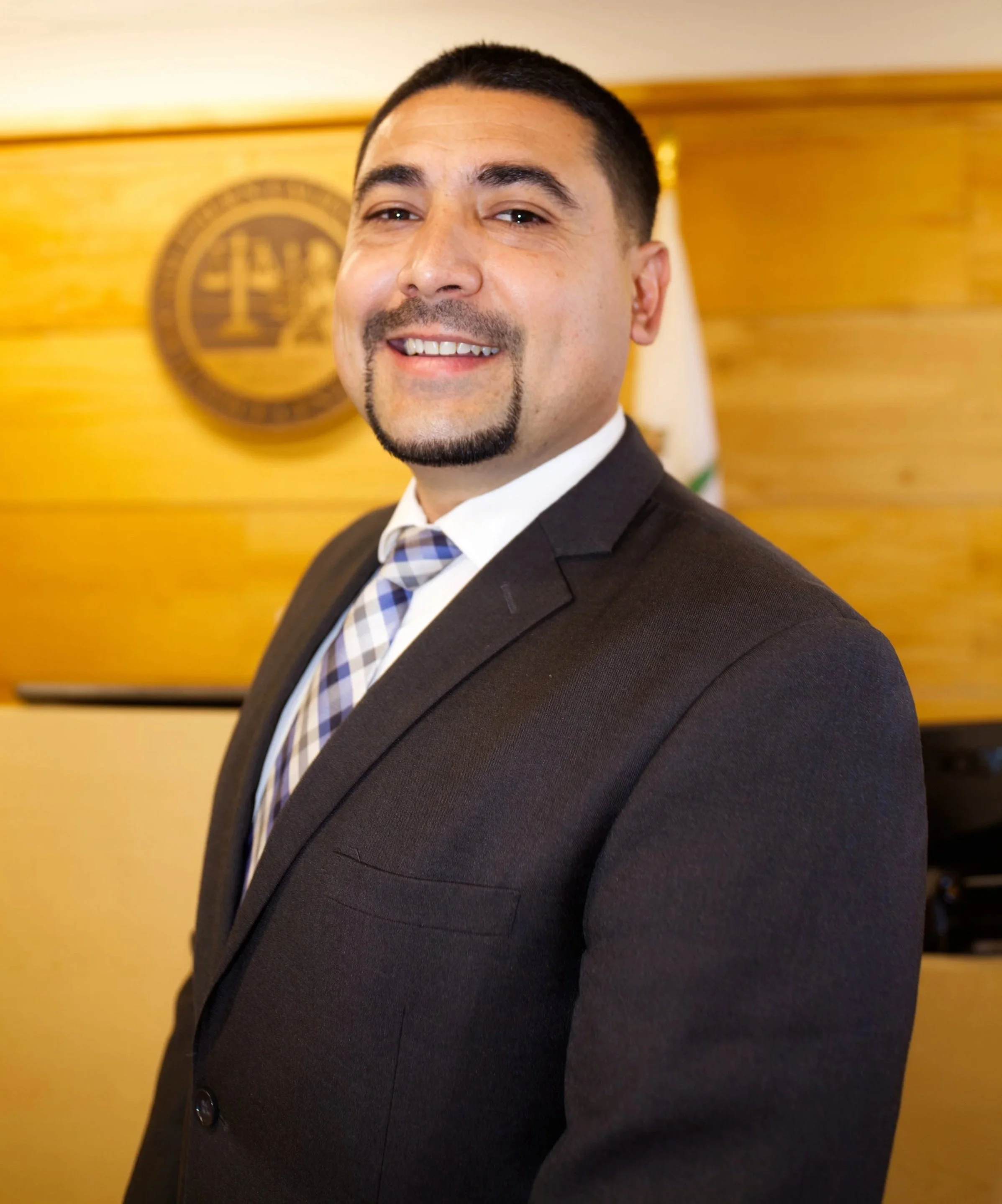 Hombre de negocios sonriendo vestido con traje en un ambiente formal, con un fondo de madera y un escudo en la pared.