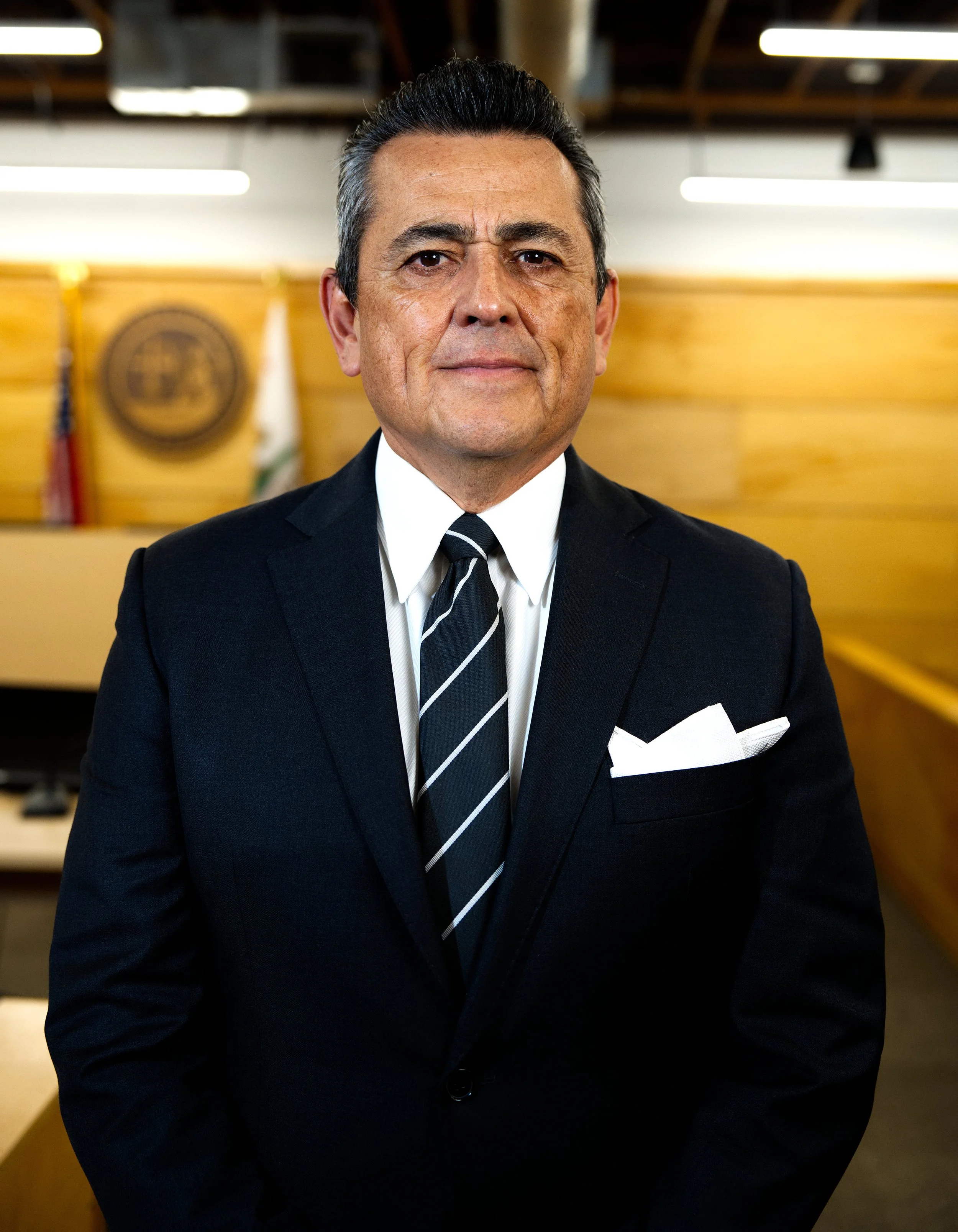Hombre con traje oscuro, camisa blanca y corbata, posando en una oficina con fondo de madera y banderas.