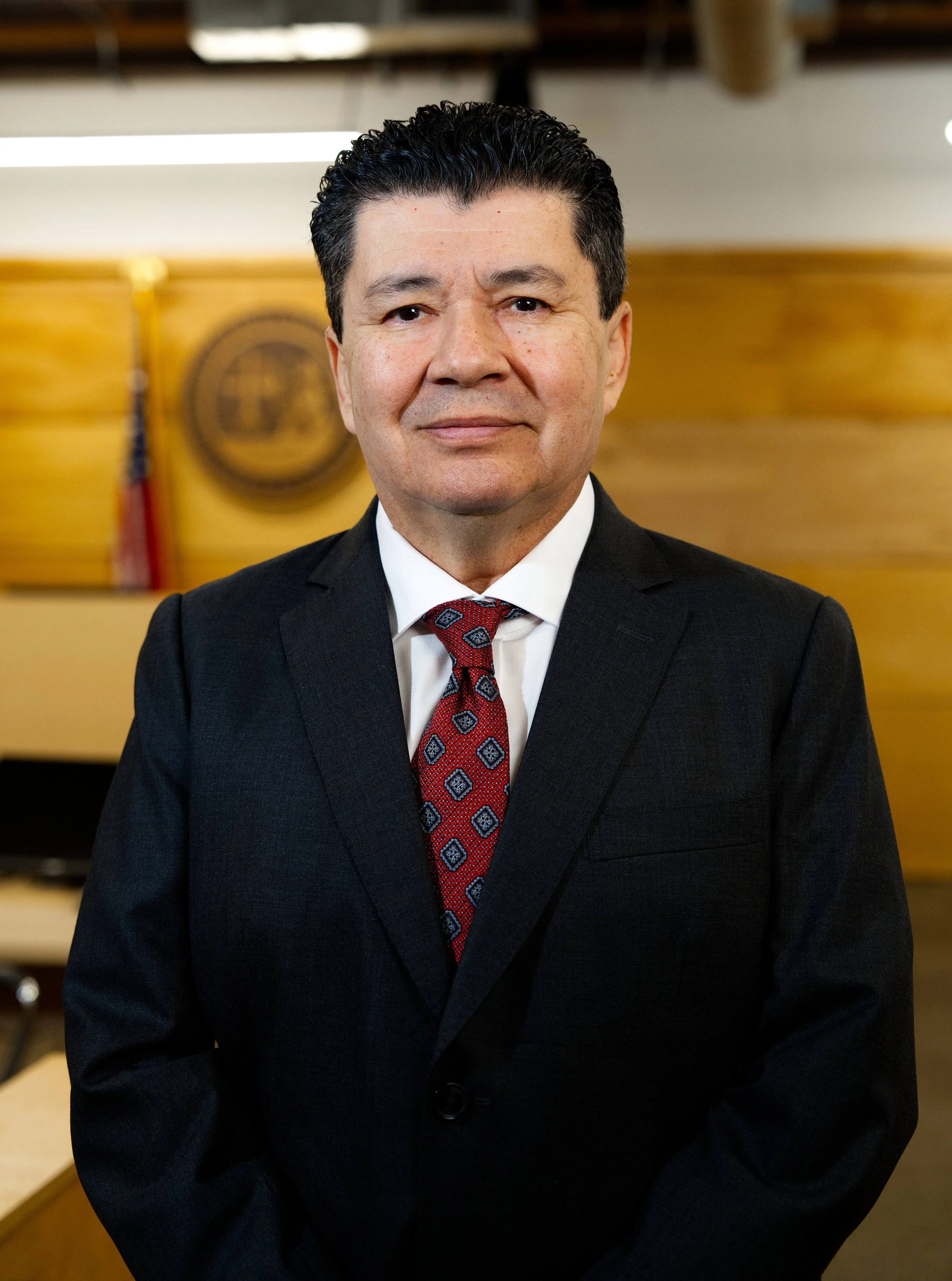 Hombre vestido con traje negro, camisa blanca y corbata de patrones, de cabello oscuro y peinado hacia atrás, en un entorno formal con fondo de madera y bandera en un fondo difuso.