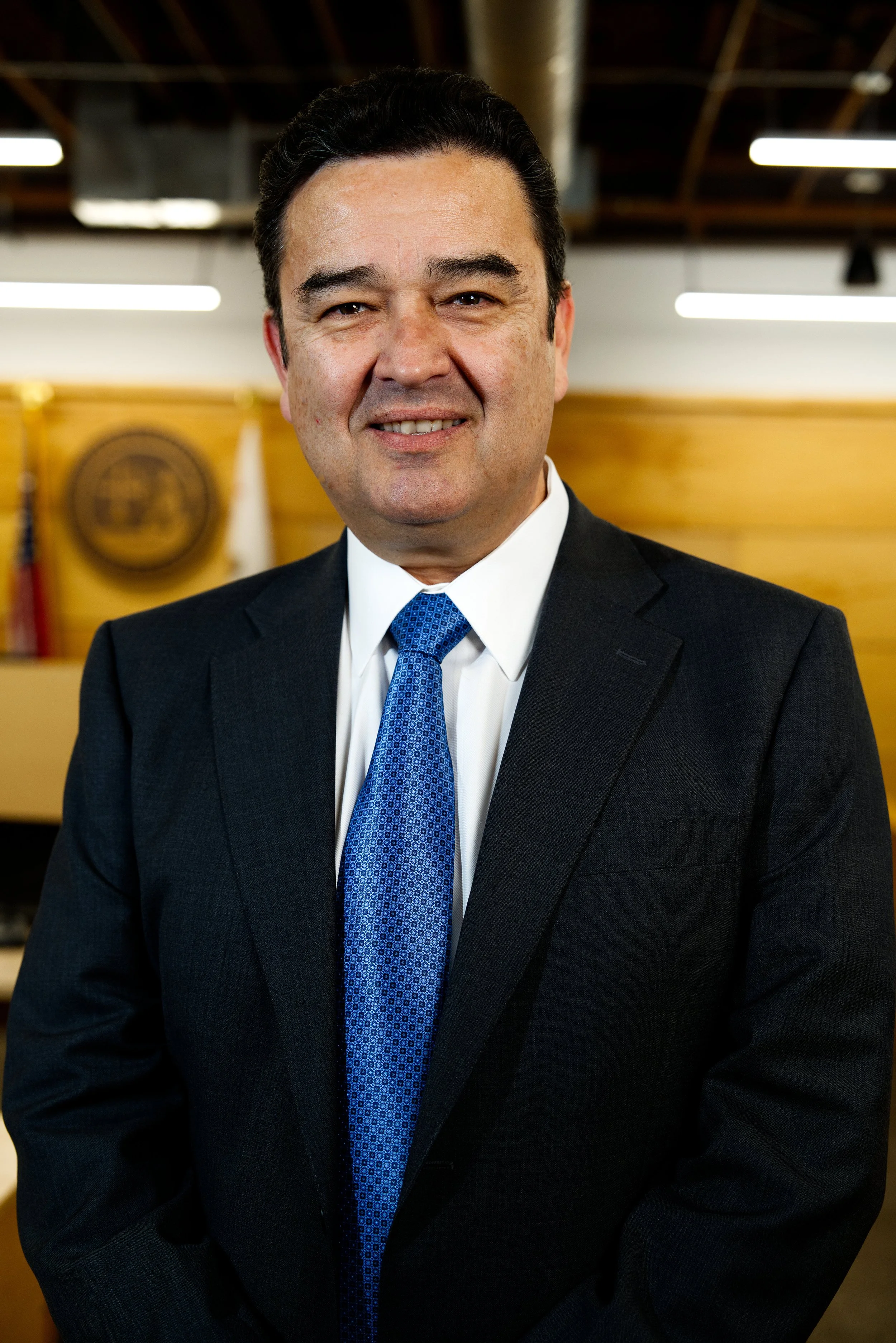 Hombre de negocios con traje negro y corbata azul, sonriendo en una oficina o conferencia.