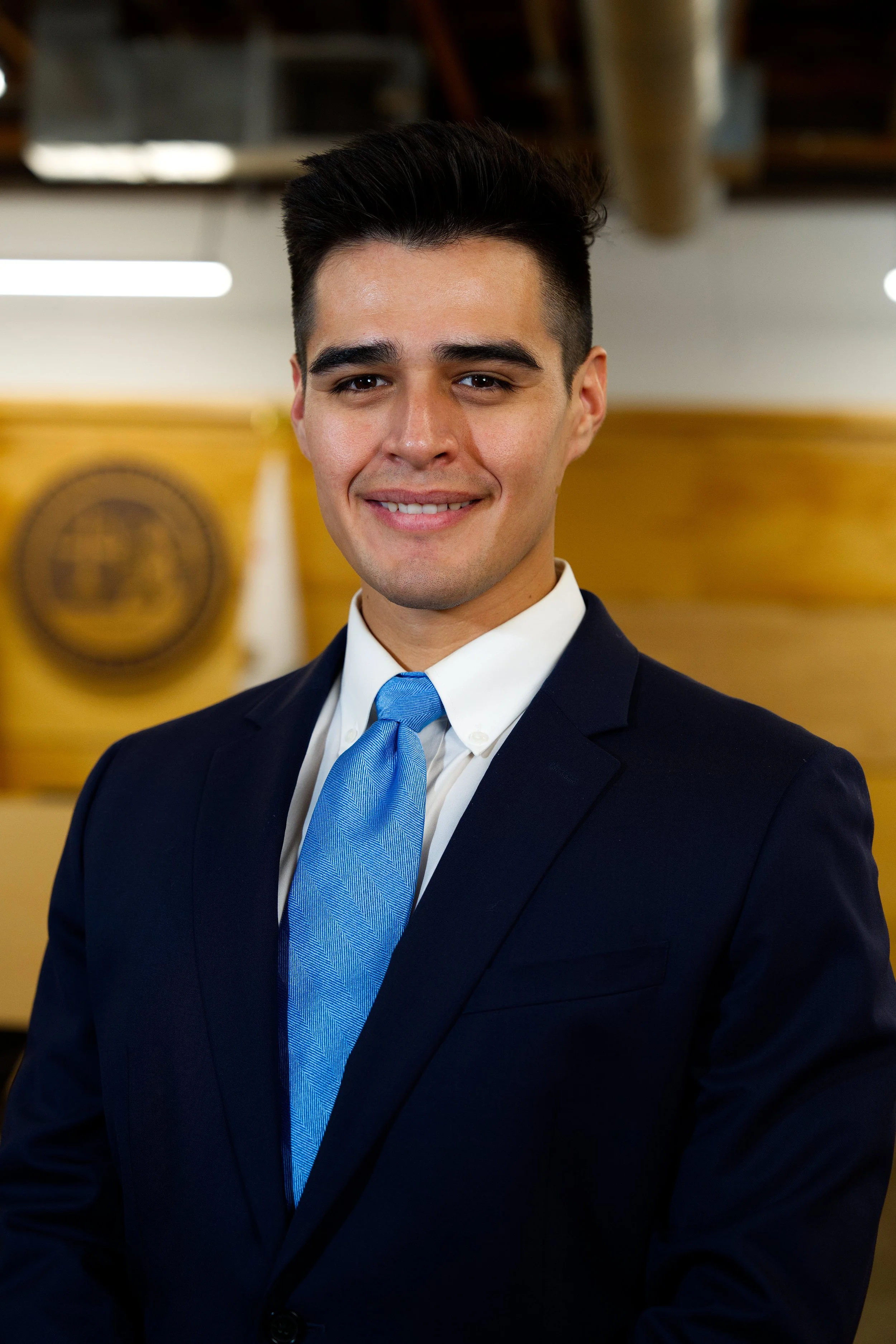 Hombre con vestimenta formal, traje oscuro y corbata azul, sonriendo en un entorno profesional.