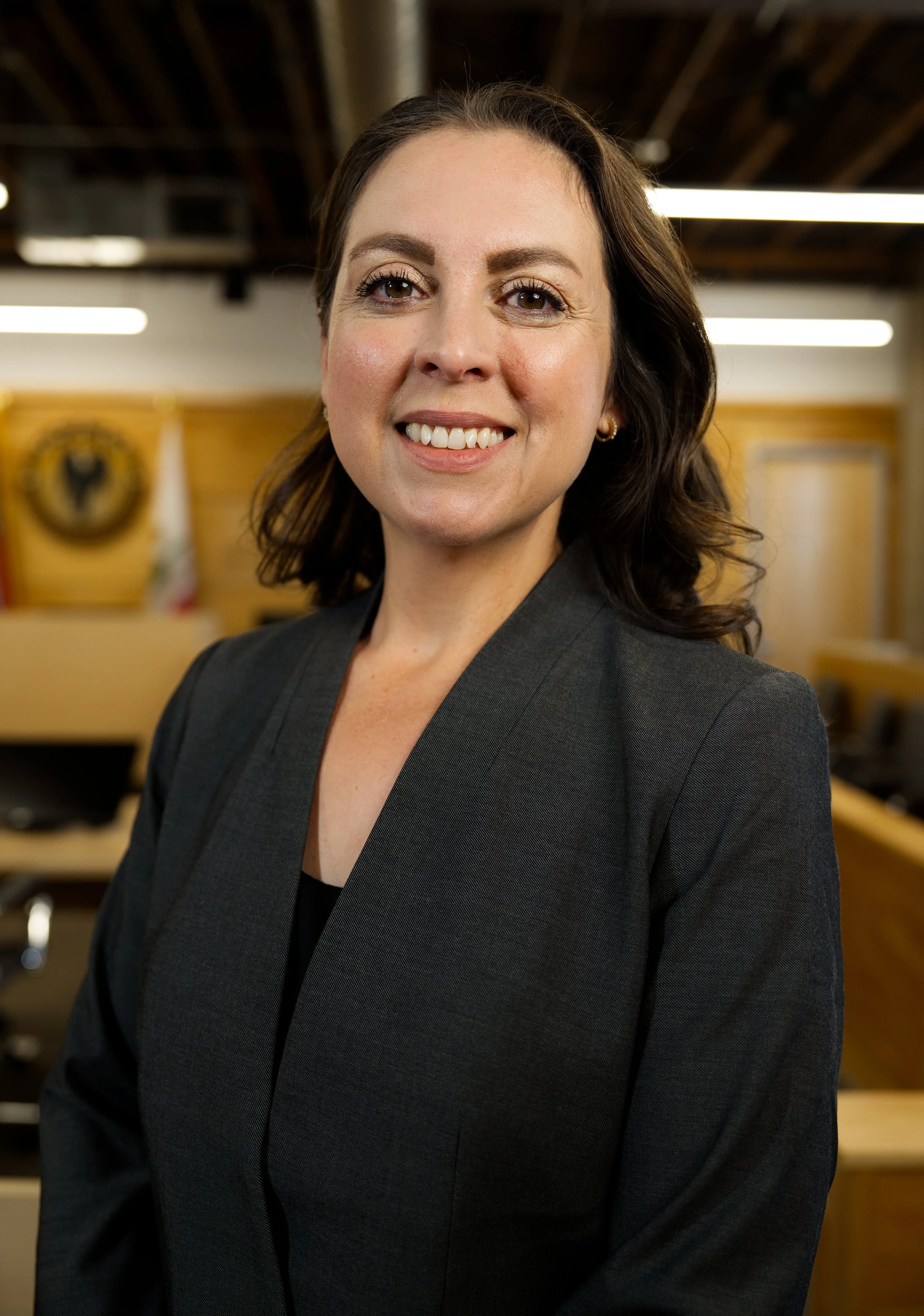 Retrato de una mujer en un entorno de oficina con fondo de madera y banderas, vistiendo una chaqueta negra y sonriendo.