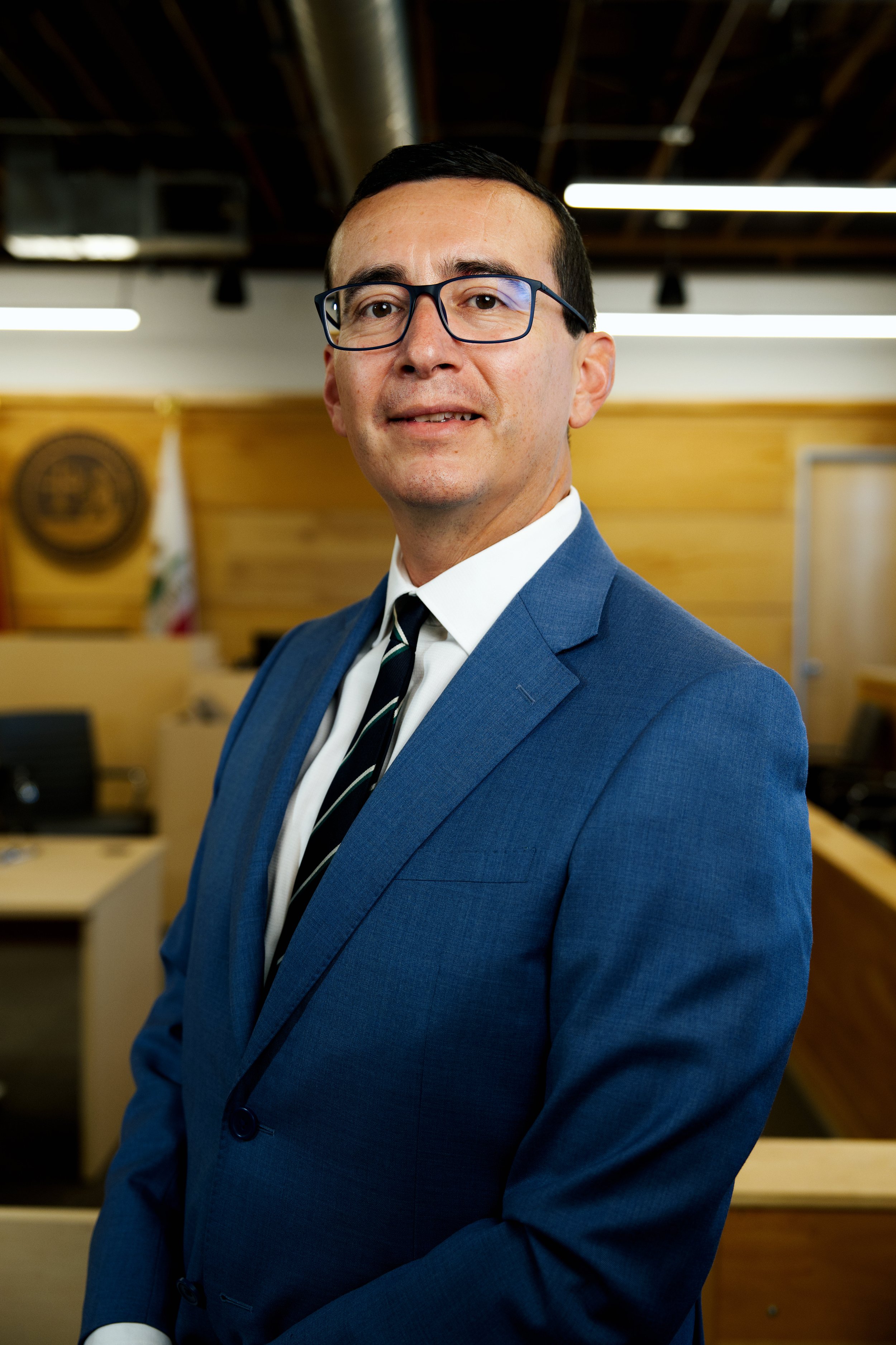 Hombre con gafas viste un traje azul, camisa blanca y corbata en una sala de juicio o reunión formal con fondo de madera y bandera de California.