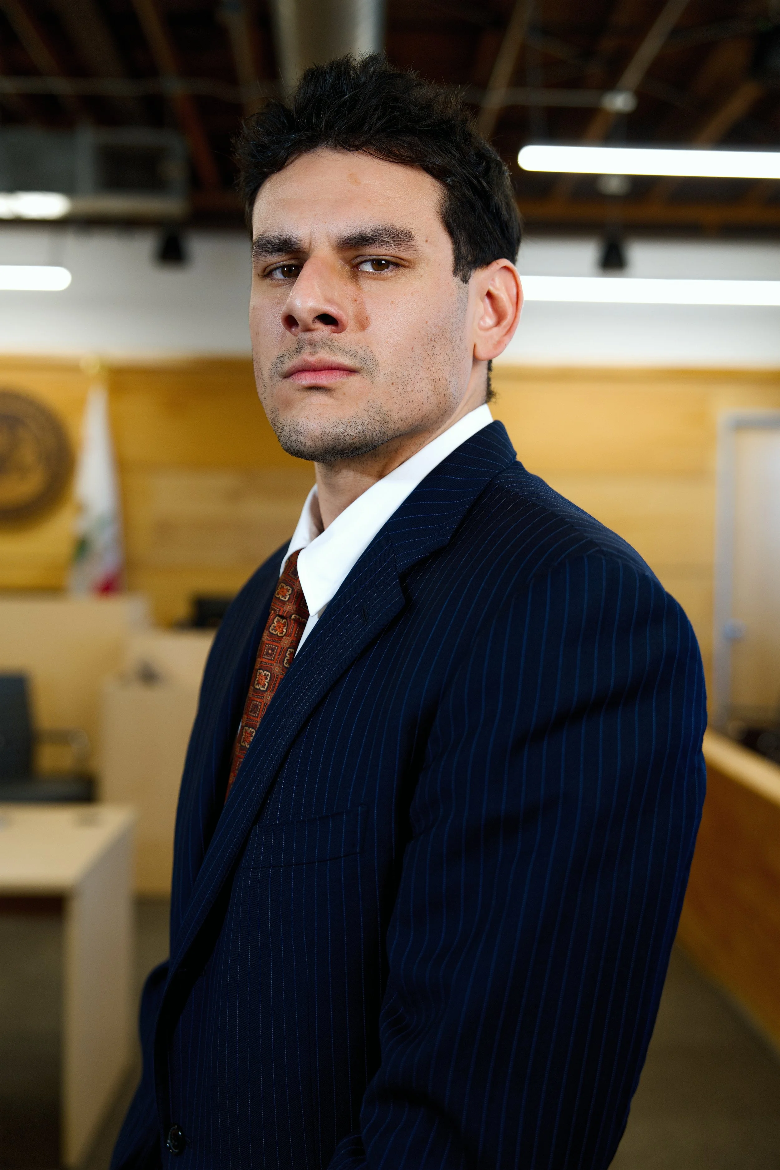 Hombre con expresión seria, vestido con traje formal y corbata, en oficina con bandera de México en el fondo.