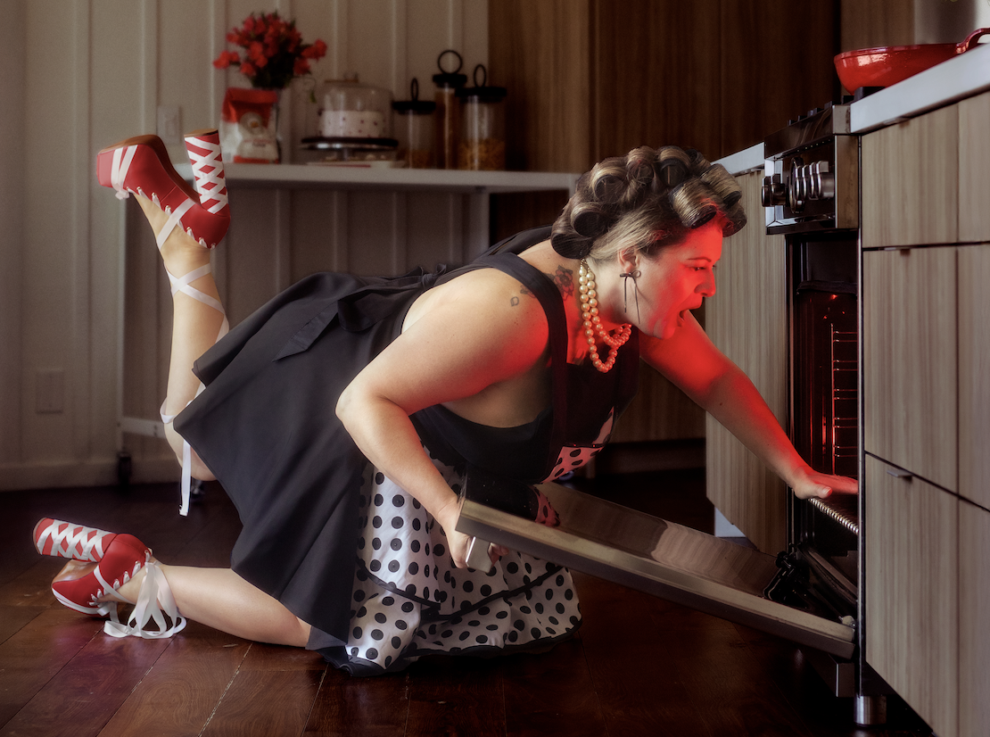 A woman dressed in vintage style with polka dot clothing and red shoes is kneeling on the floor and opening an oven, with her head inside the oven.