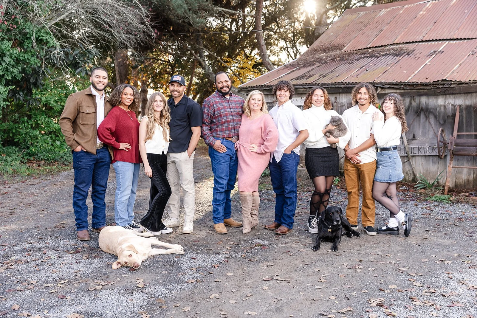 Group of people standing outdoors in front of an old barn with trees in the background, posing for a photo with two dogs, one lying on the ground and one being held, and a small skateboard.