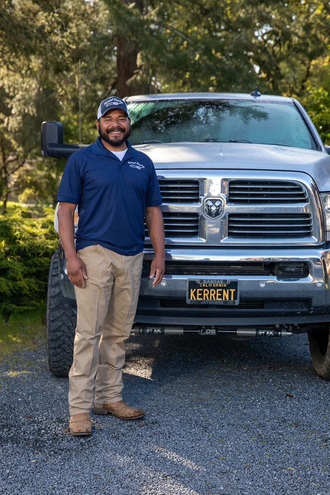 owner of Kerrent land services wearing a blue polo shirt, beige pants, and boots, in front of a large silver Ram pickup truck with a license plate that reads 'KERRent', with trees and green foliage in the background in Sebastopol Northern California.