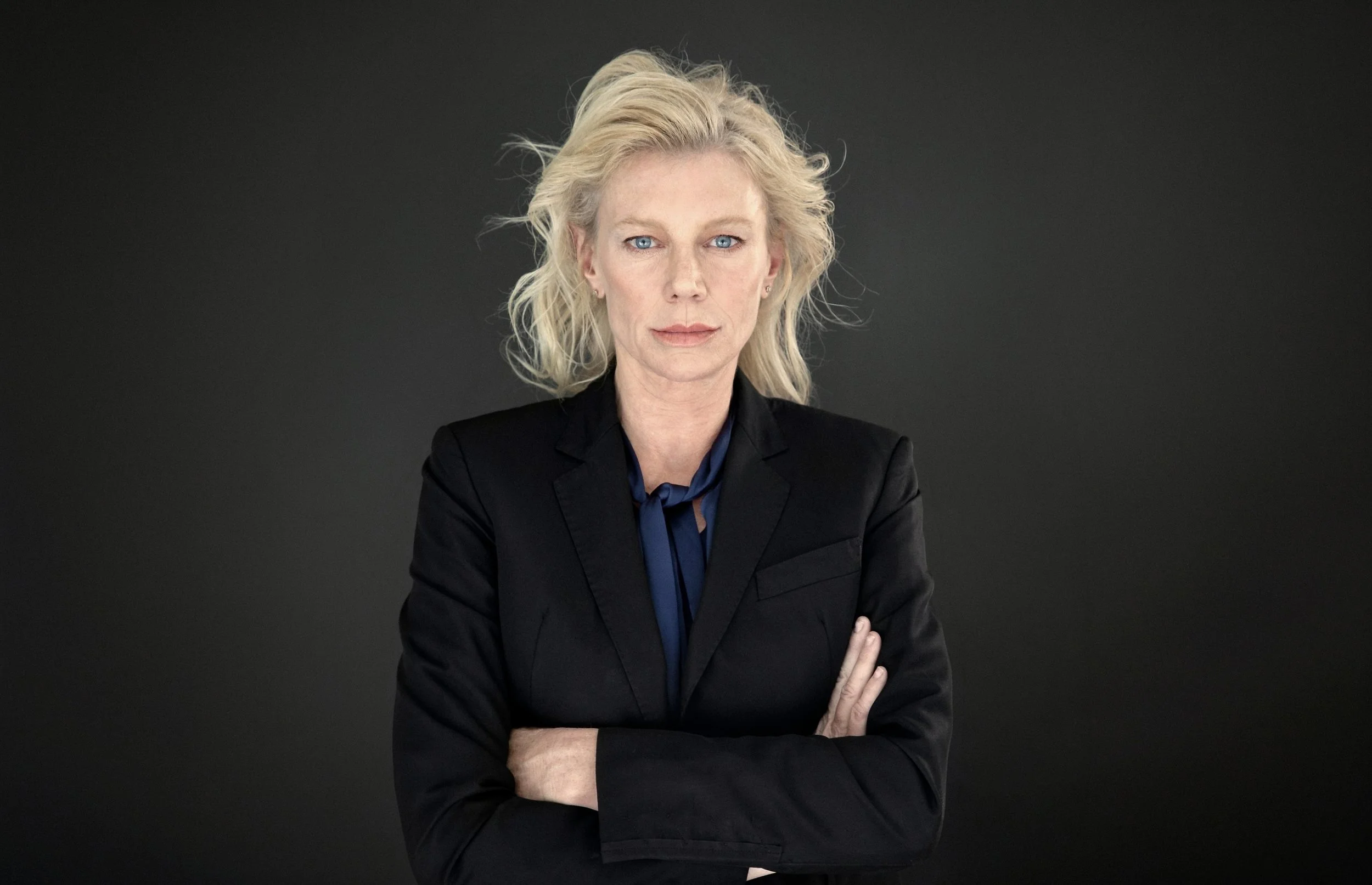 Portrait of Peta Wilson wearing a black suit against a dark background.