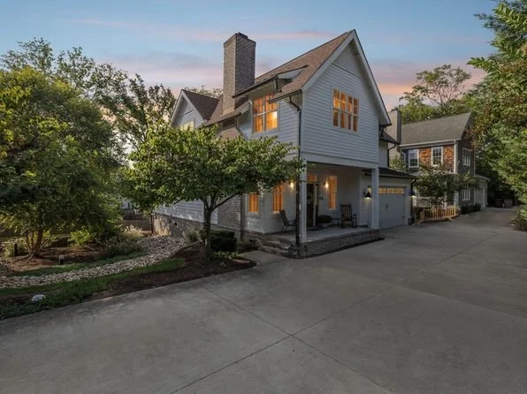 A two-story house with a gabled roof, white siding, and lit windows, surrounded by trees and greenery, with a driveway and a neighboring house visible.