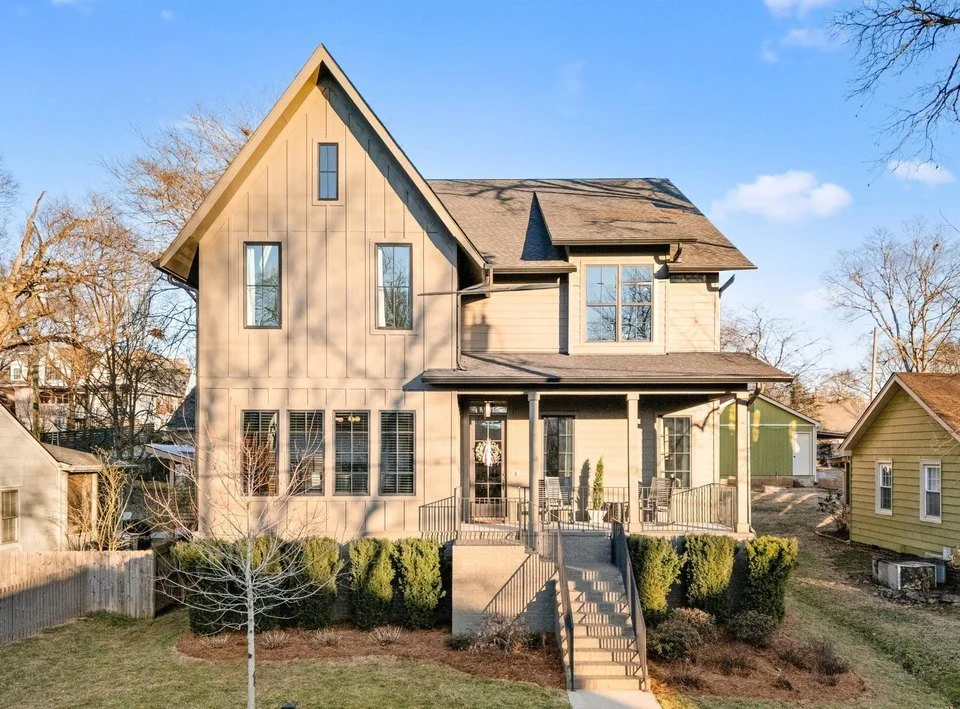 Large two-story house with beige siding, gabled roof, and front porch, surrounded by trees and neighboring houses under a blue sky.