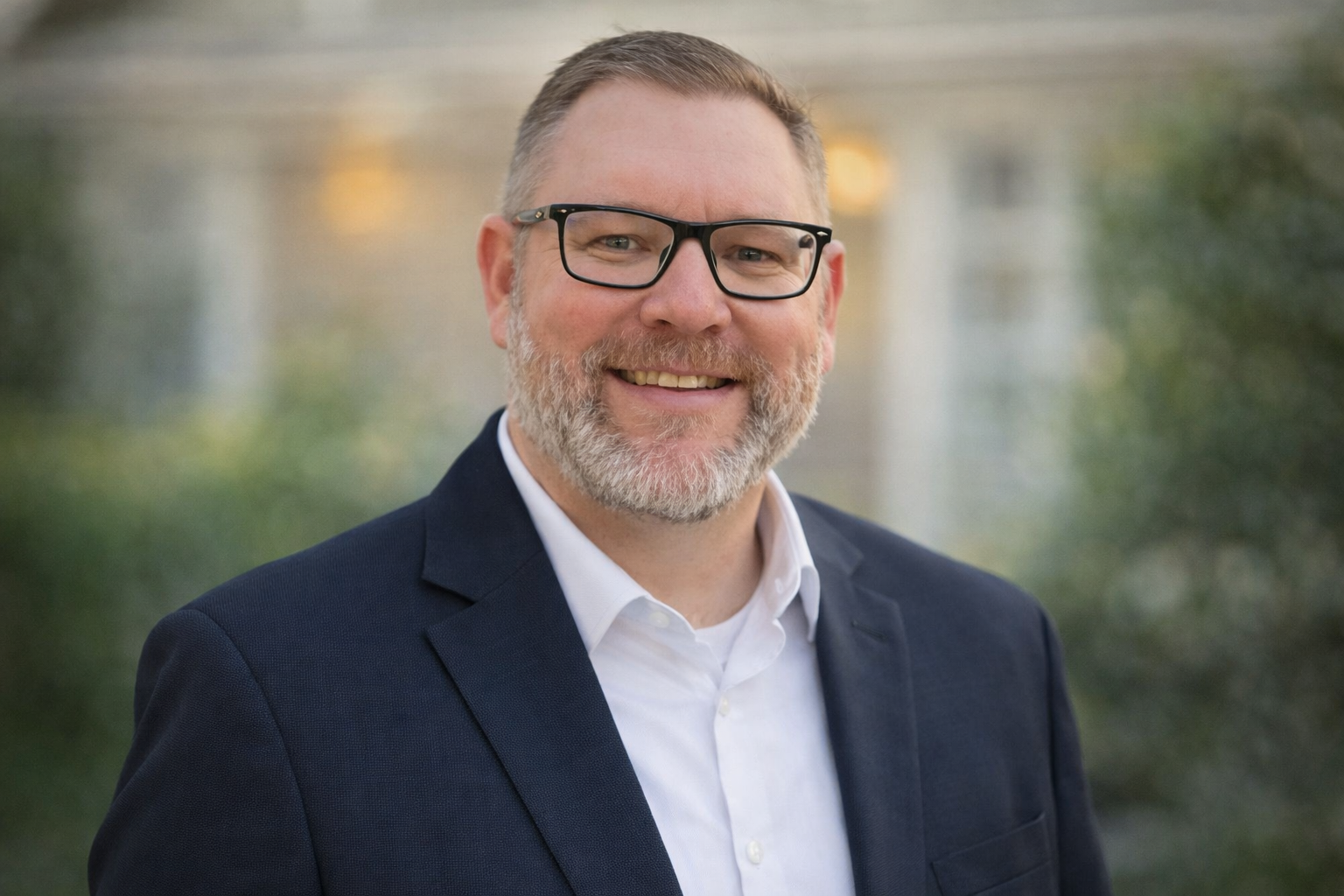 A middle-aged man with glasses, a beard, and short hair, dressed in a suit jacket and white shirt, smiling outdoors with blurred greenery in the background.