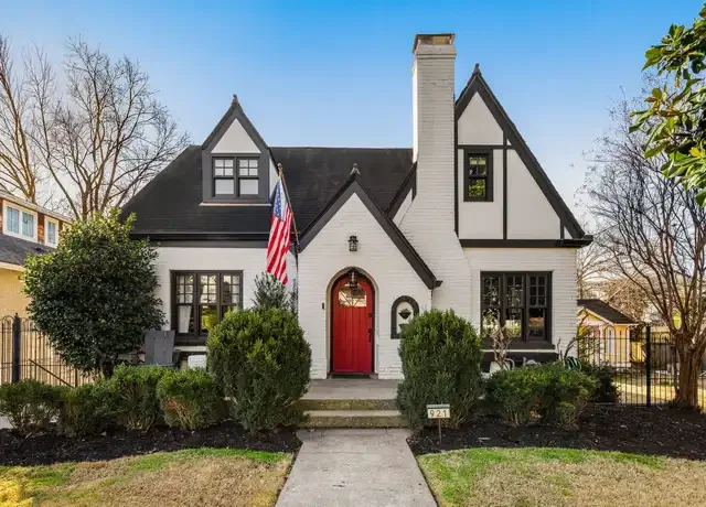 Front view of a white house with black details, a red front door, and an American flag, surrounded by a well-maintained lawn and bushes, under a clear blue sky.