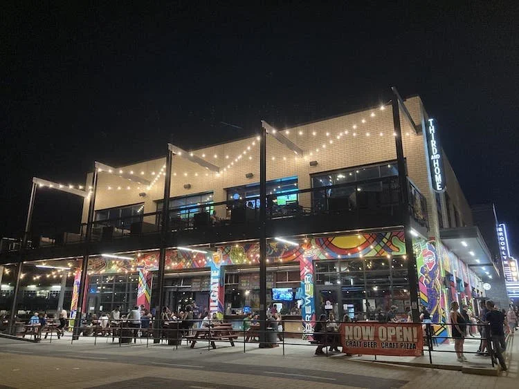 A two-story restaurant or bar with colorful exterior murals, outdoor seating, and string lights at night.