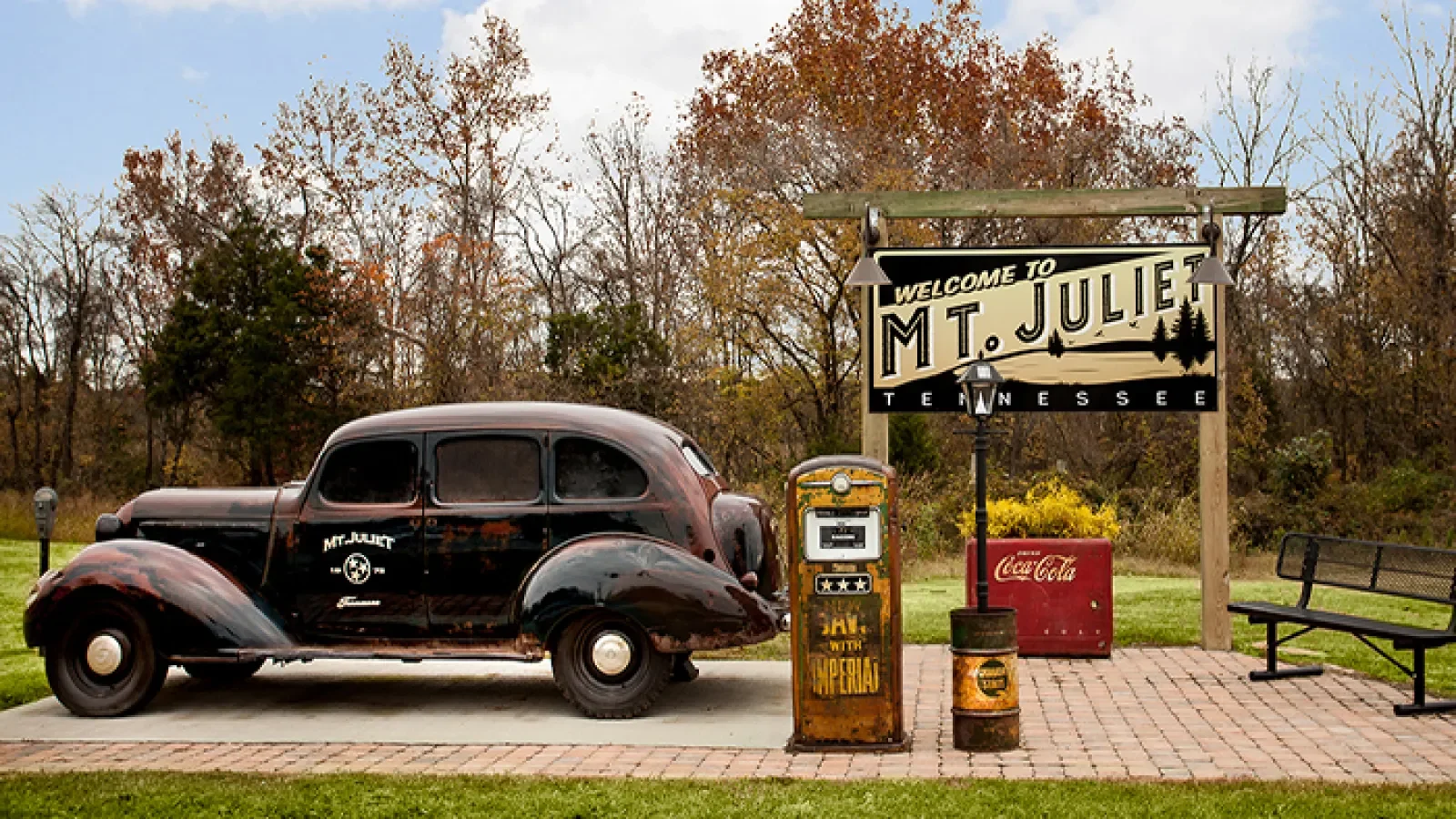 Vintage black car parked beside a historic sign welcoming visitors to Mount Juliet, Tennessee, with an old gas pump, a Coca-Cola cooler, and a bench nearby, set against autumn trees.