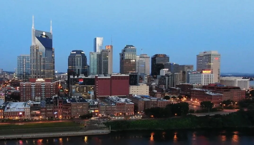 Skyline of Nashville with various skyscrapers and buildings, a river in the foreground, and city lights beginning to turn on at dusk.