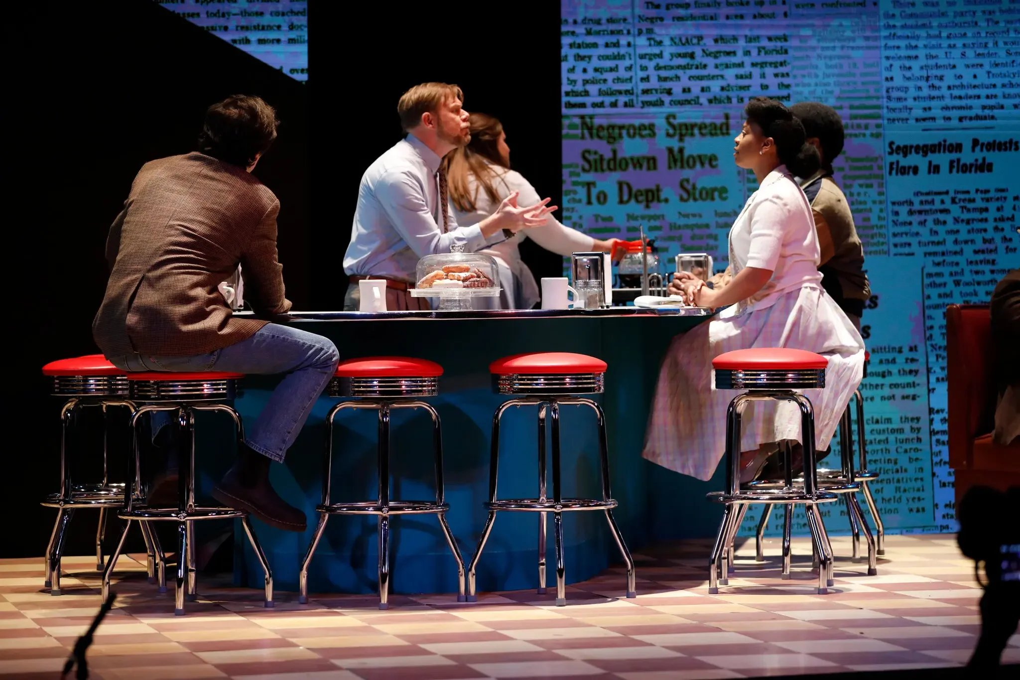 A scene from a theatrical production depicting a period setting with five actors standing behind a bar counter, engaged in a conversation. The background features a large, colorful newspaper print projection with headlines related to segregation and social issues.