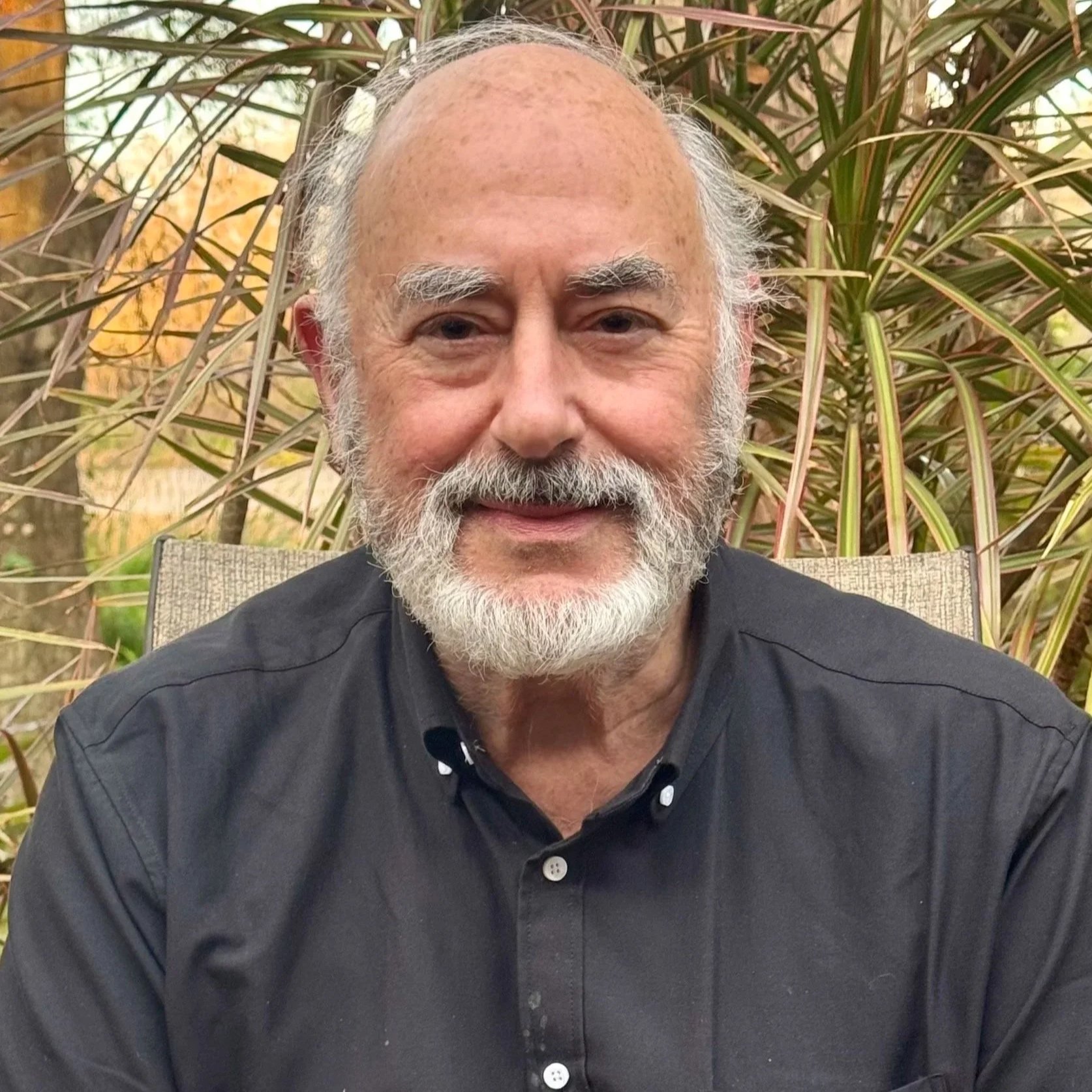A smiling elderly man with a beard, wearing a black shirt, sitting outdoors in front of green foliage.